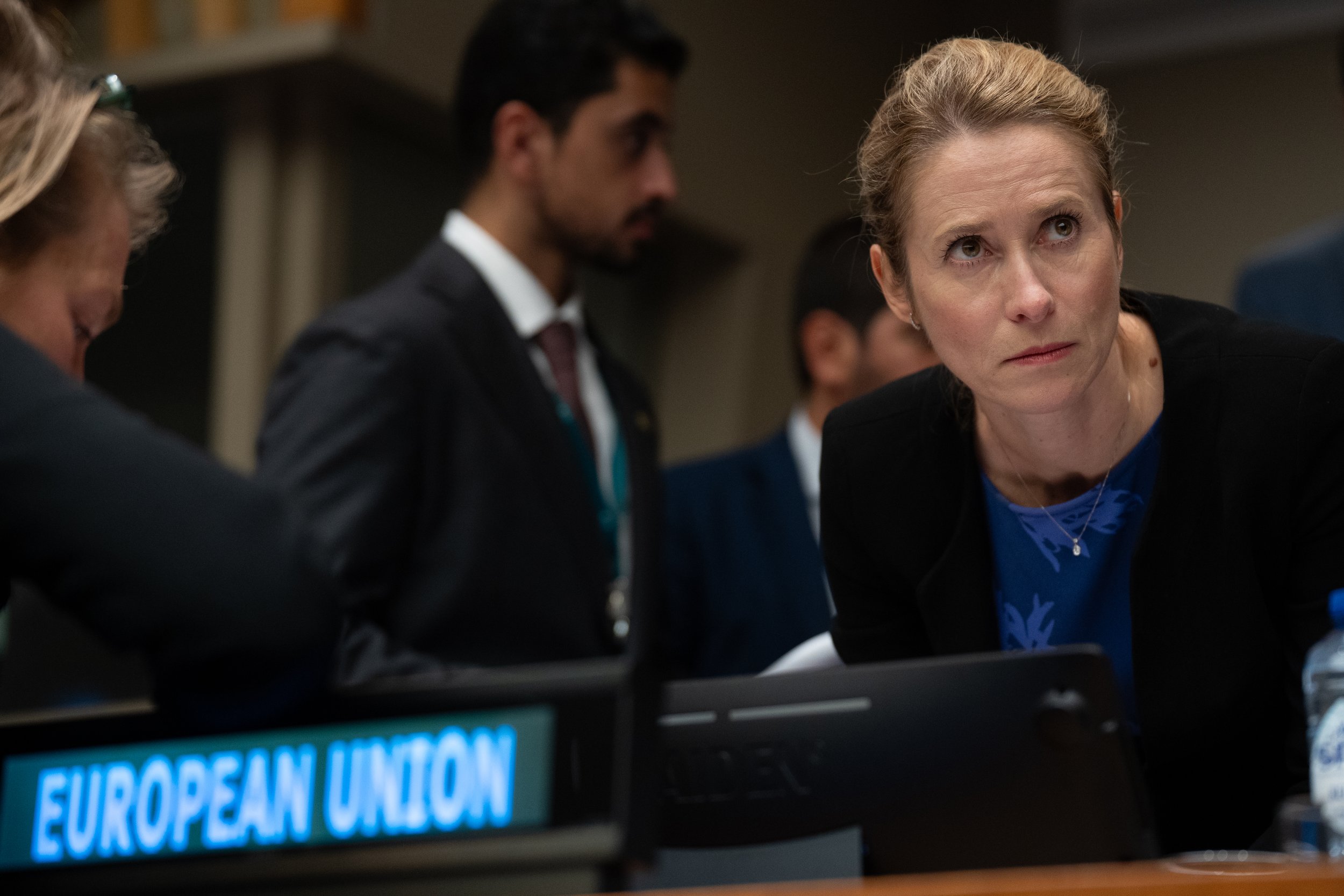 A woman with blonde hair in a bun, wearing a black blazer and blue top, leaning forward at a conference table with a sign that says 'European Union', listening intently.