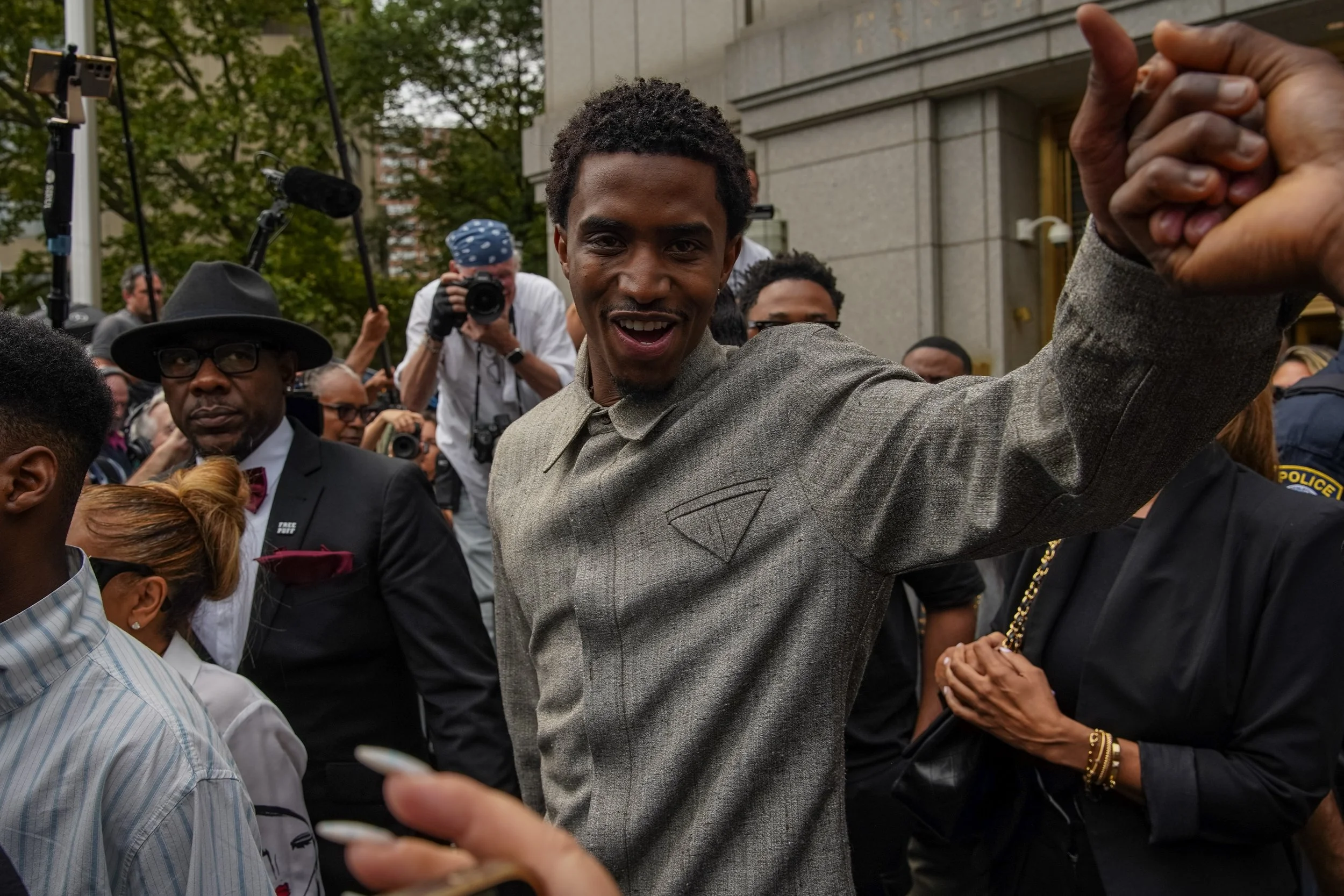 Man smiling and raising his fist in a crowd of people outside a building, surrounded by photographers and onlookers.