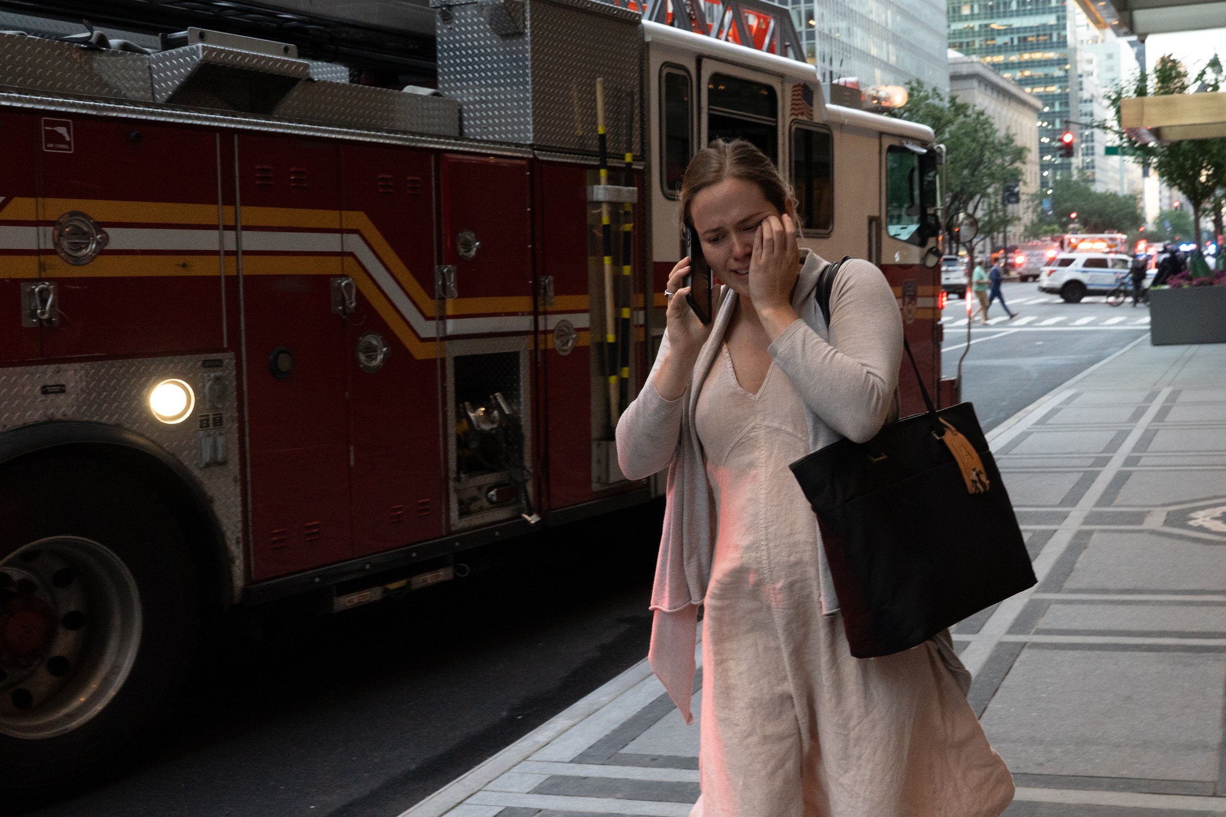 A woman with a distressed expression talking on her cell phone, standing on a city sidewalk near a fire truck, with traffic and tall buildings in the background.
