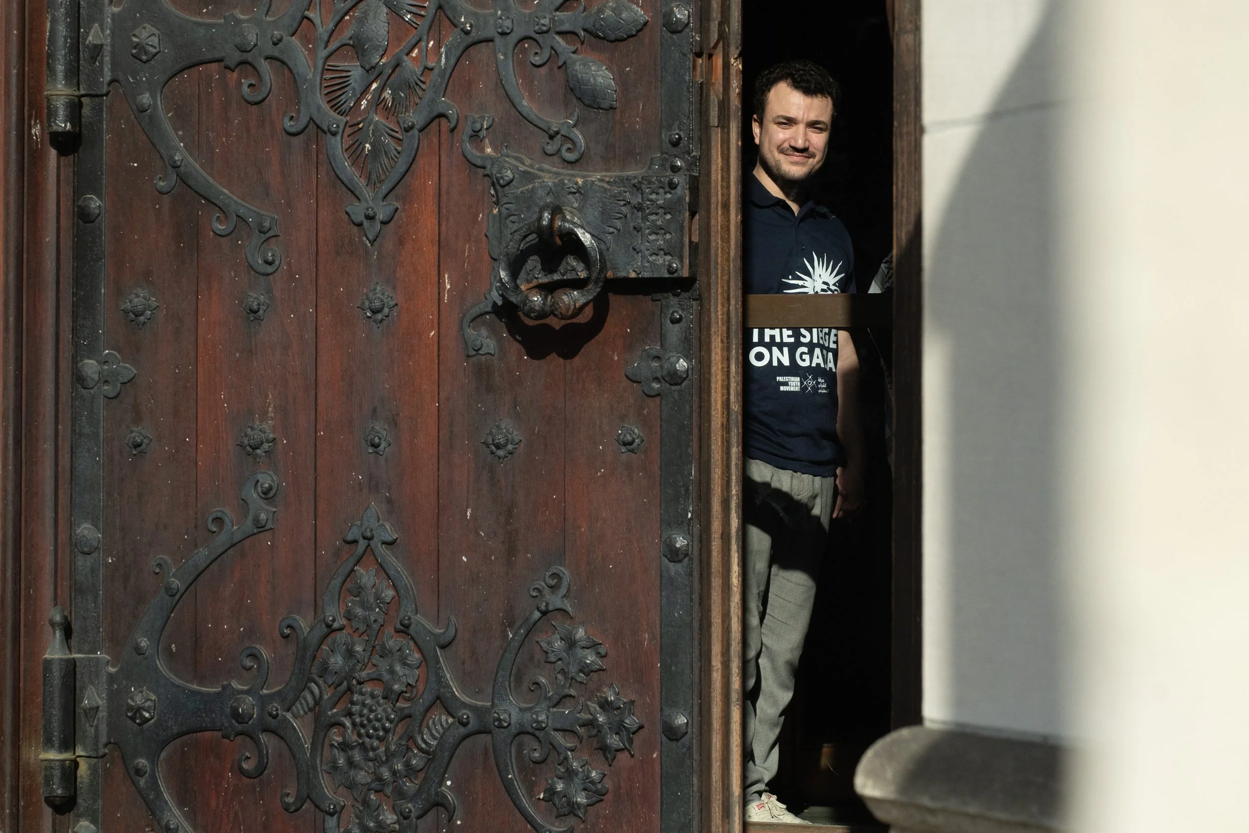 A man peeking through a partially open wooden door with ornate metalwork, standing inside a building, with sunlight illuminating part of the scene.