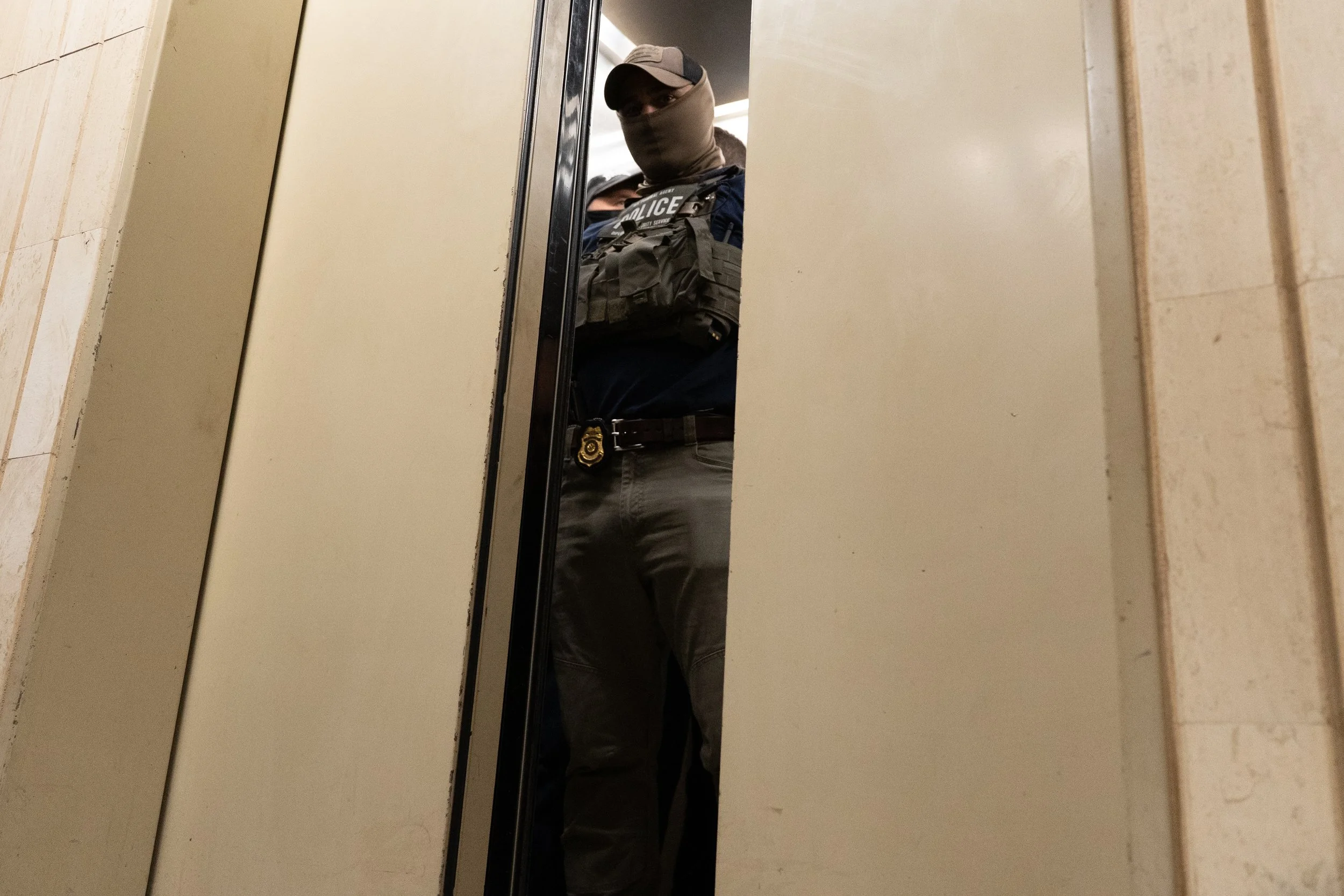A police officer standing in front of an elevator door, wearing tactical gear and a face covering, with only part of their body visible through the partially open elevator door.