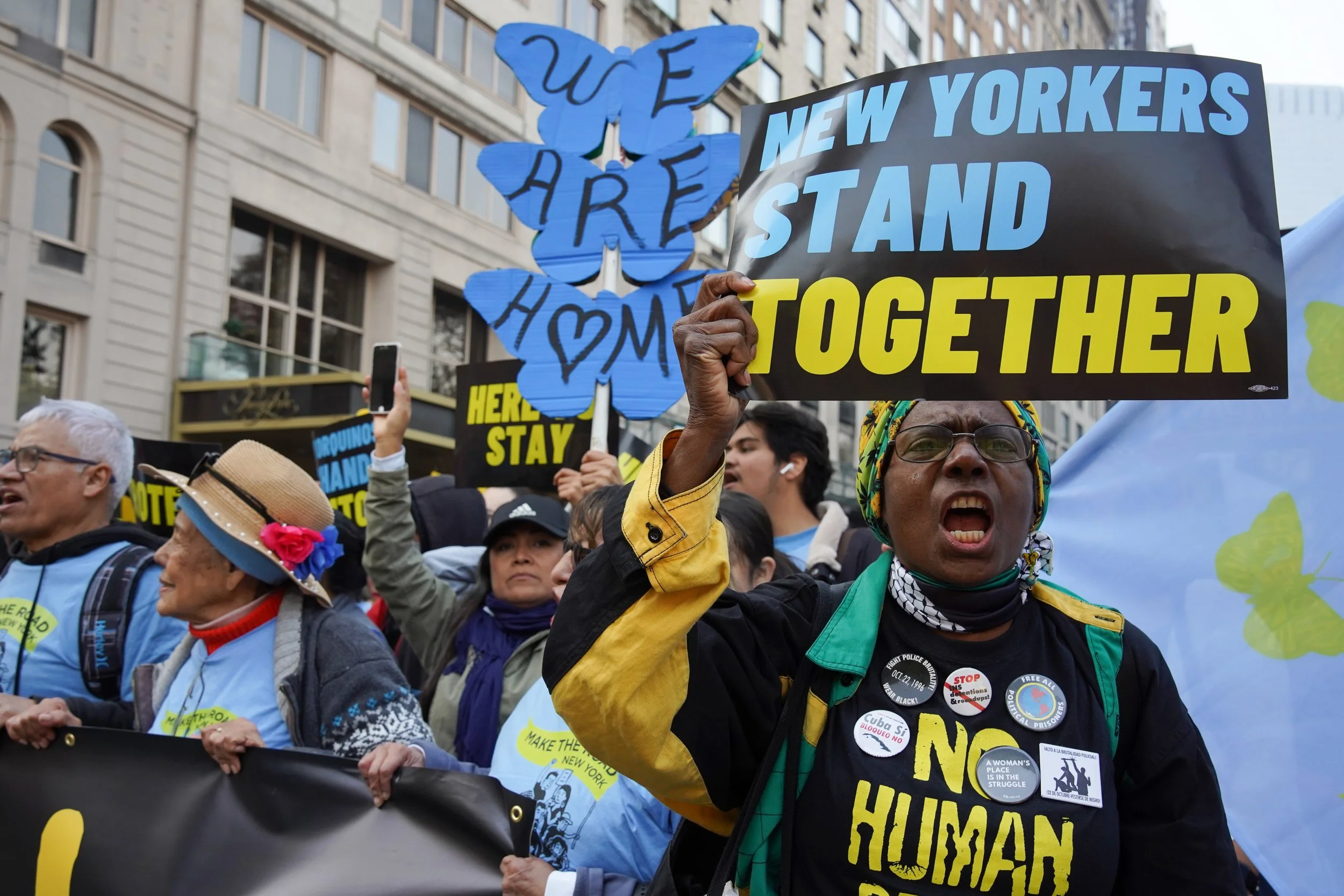 Protesters in a street rally holding signs with messages about unity and human rights, including a woman with glasses and a headscarf yelling, a woman with a hat and roses, and other people with signs and banners in the background.