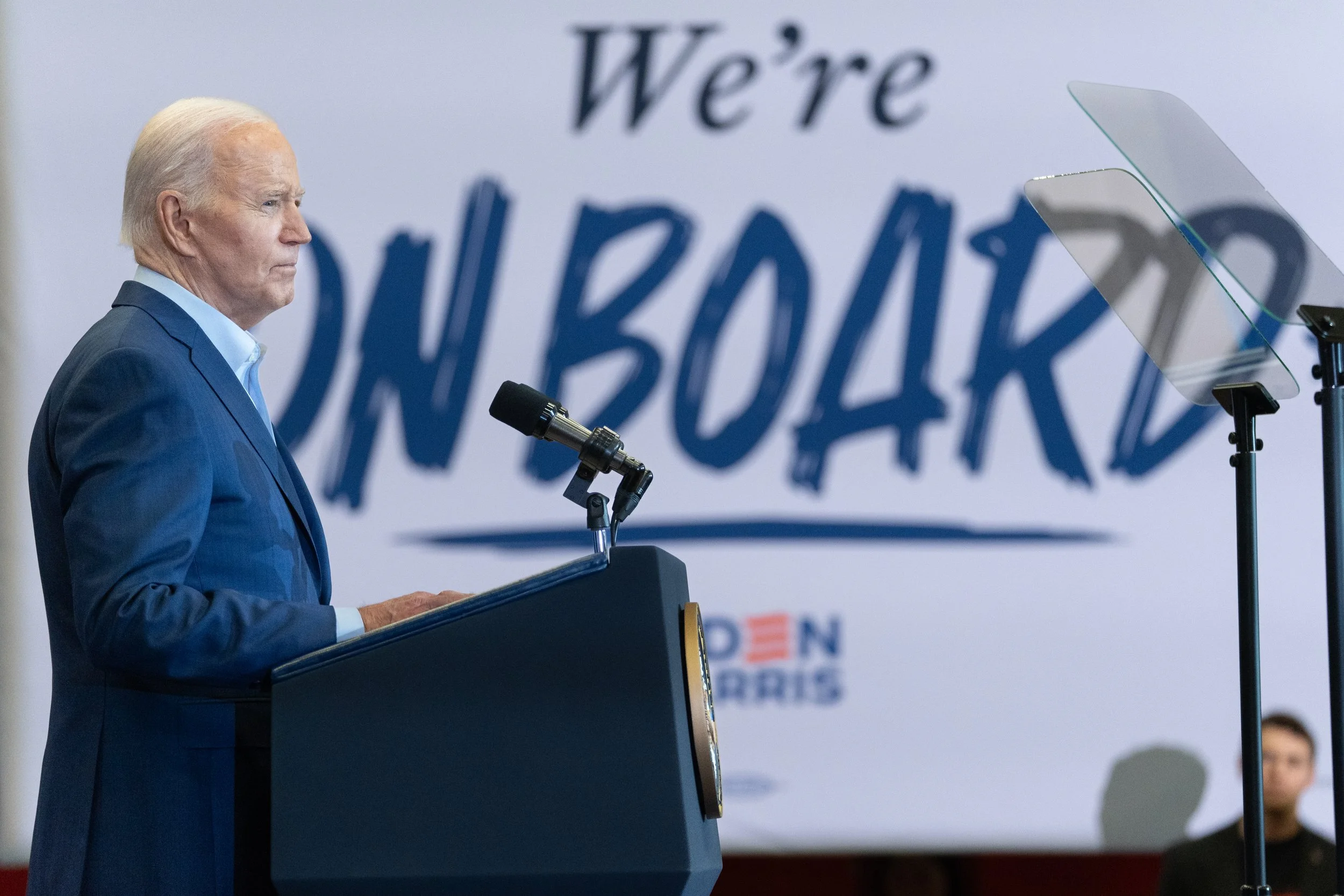 Joe Biden standing at a podium during a campaign event, with a large banner behind him that says "We're ON BOARD" and a logo for his 2024 presidential campaign.