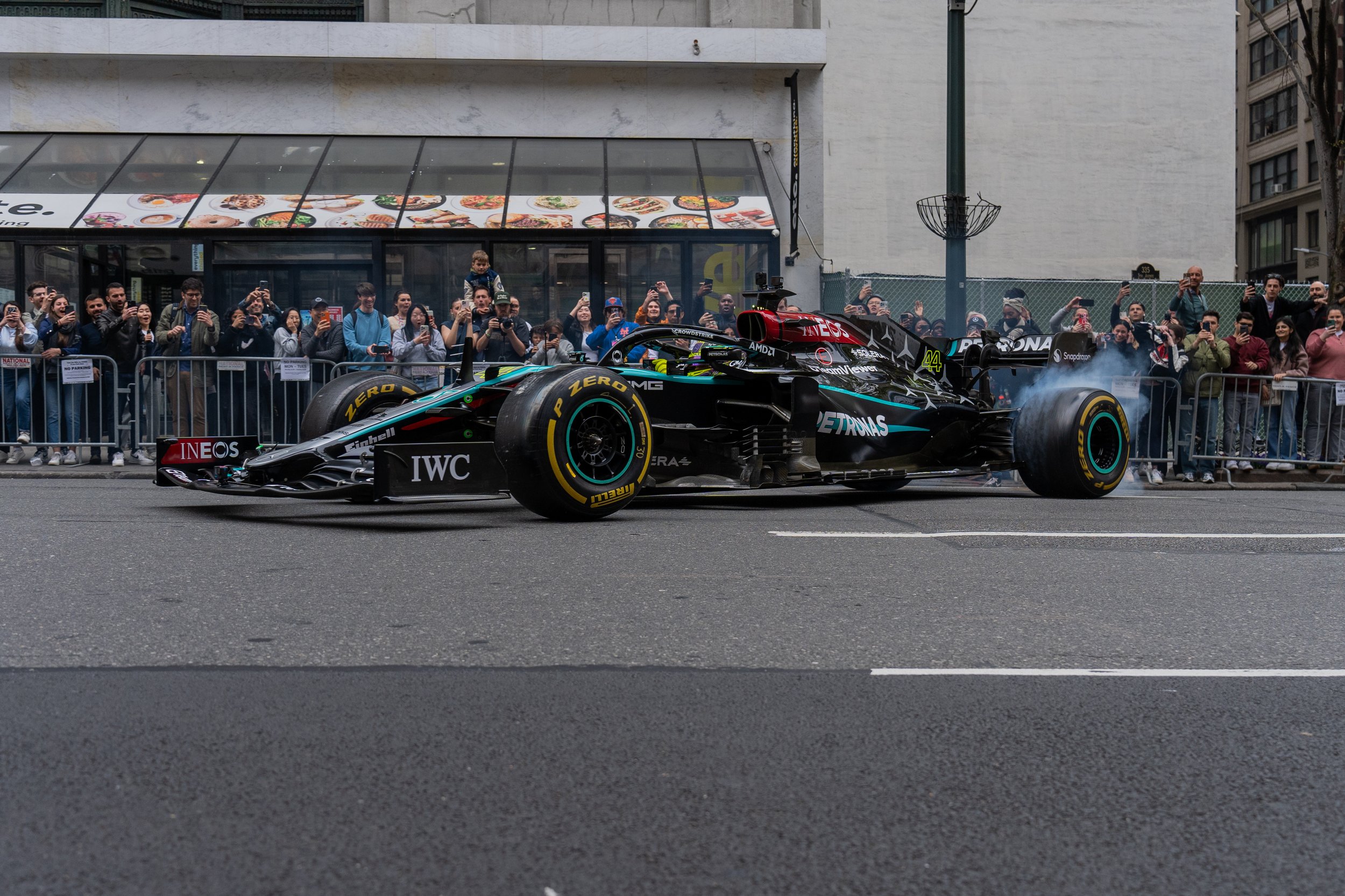 A black Formula 1 race car driving on the street during a parade or event, surrounded by onlookers behind barriers taking photos, in an urban area with buildings.