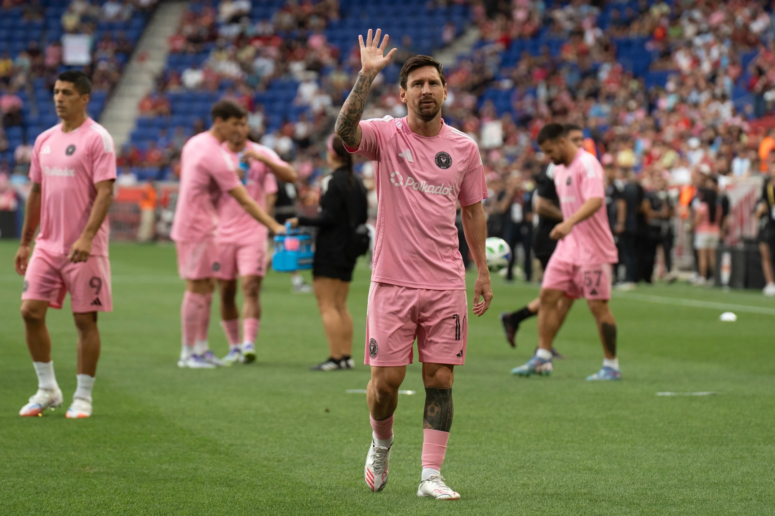 Soccer player in pink uniform waving to crowd on field, teammates and spectators in background