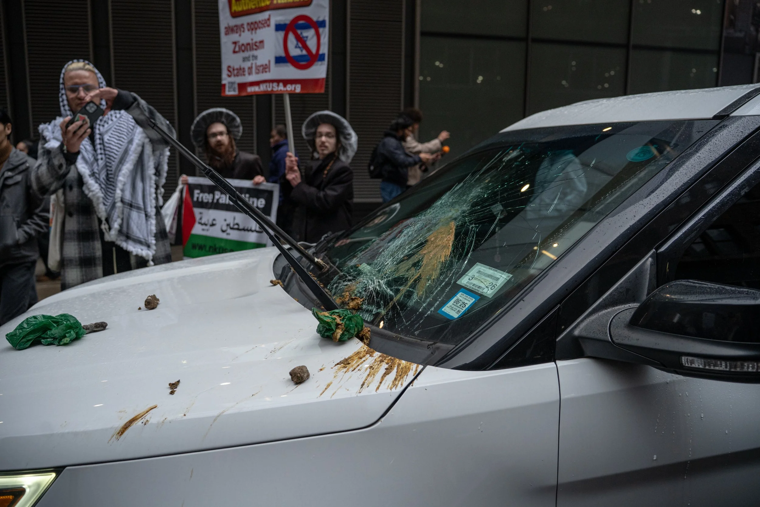 A white vehicle with a shattered windshield and damage to the hood, surrounded by mud and debris, with a group of protesters in the background holding signs, one of which opposes Zionism and Israel, at a protest march.