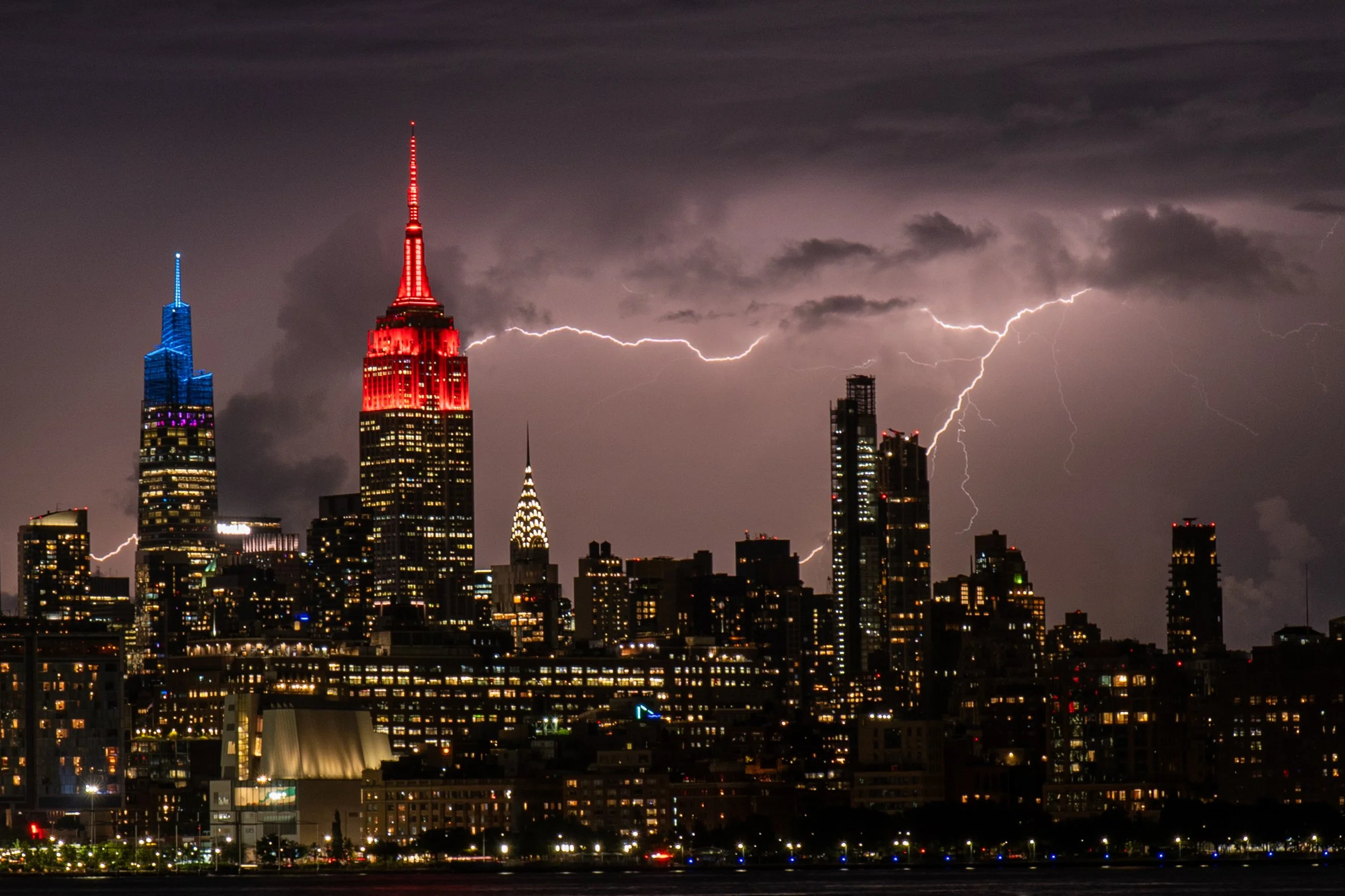 Nighttime city skyline with tall buildings illuminated, dark clouds, and multiple lightning bolts striking in the sky.