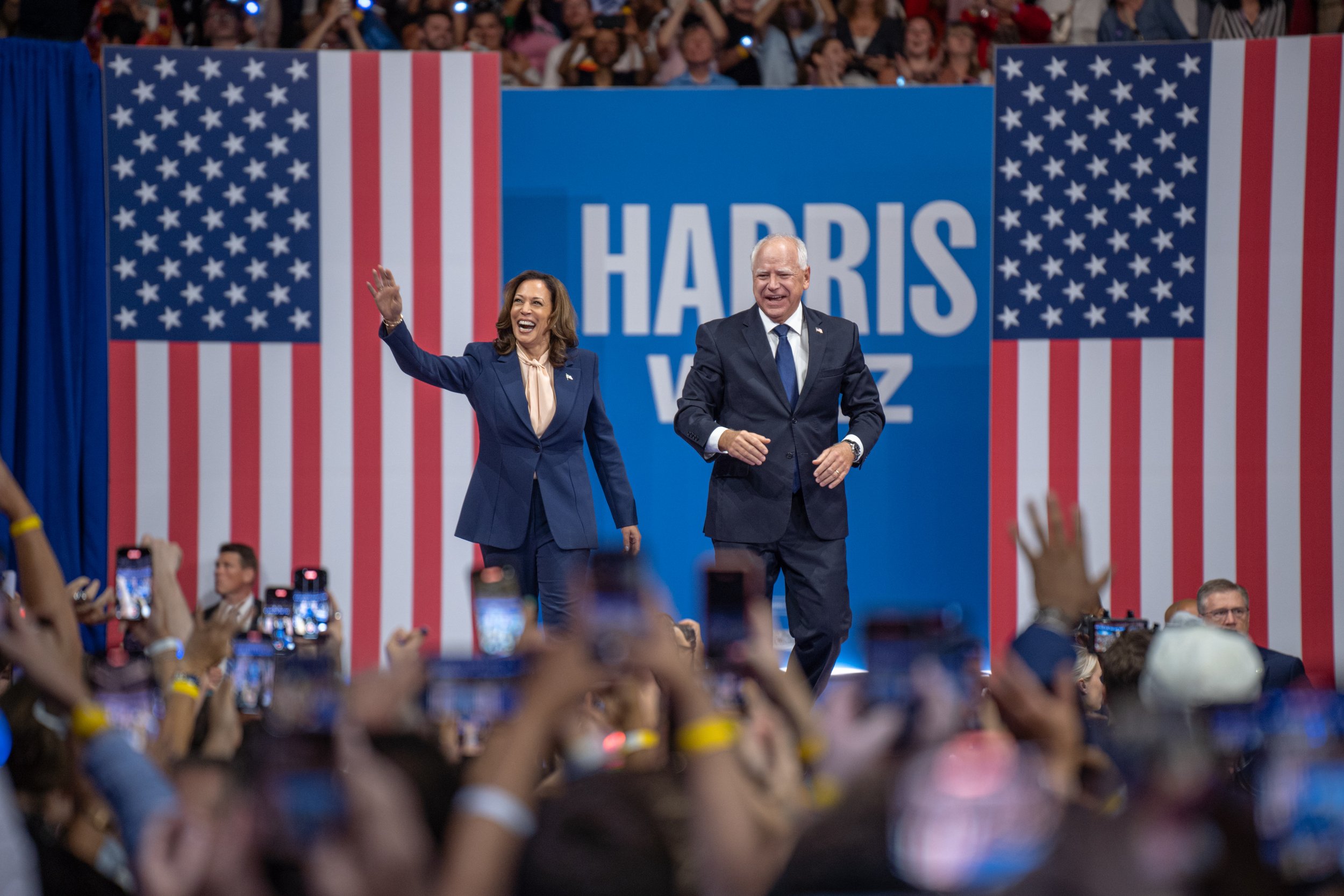 Kamala Harris and Joe Biden on stage during a political event with large American flags and a sign that says "HARRIS V. Z" in the background, surrounded by an audience taking photos and waving.