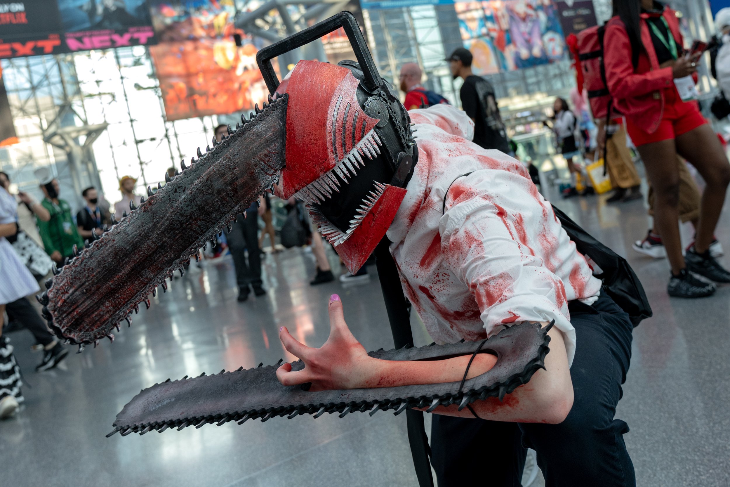 Person dressed in a horror-themed costume with a large chainsaw headpiece and bloodstained clothes at a crowded convention or event