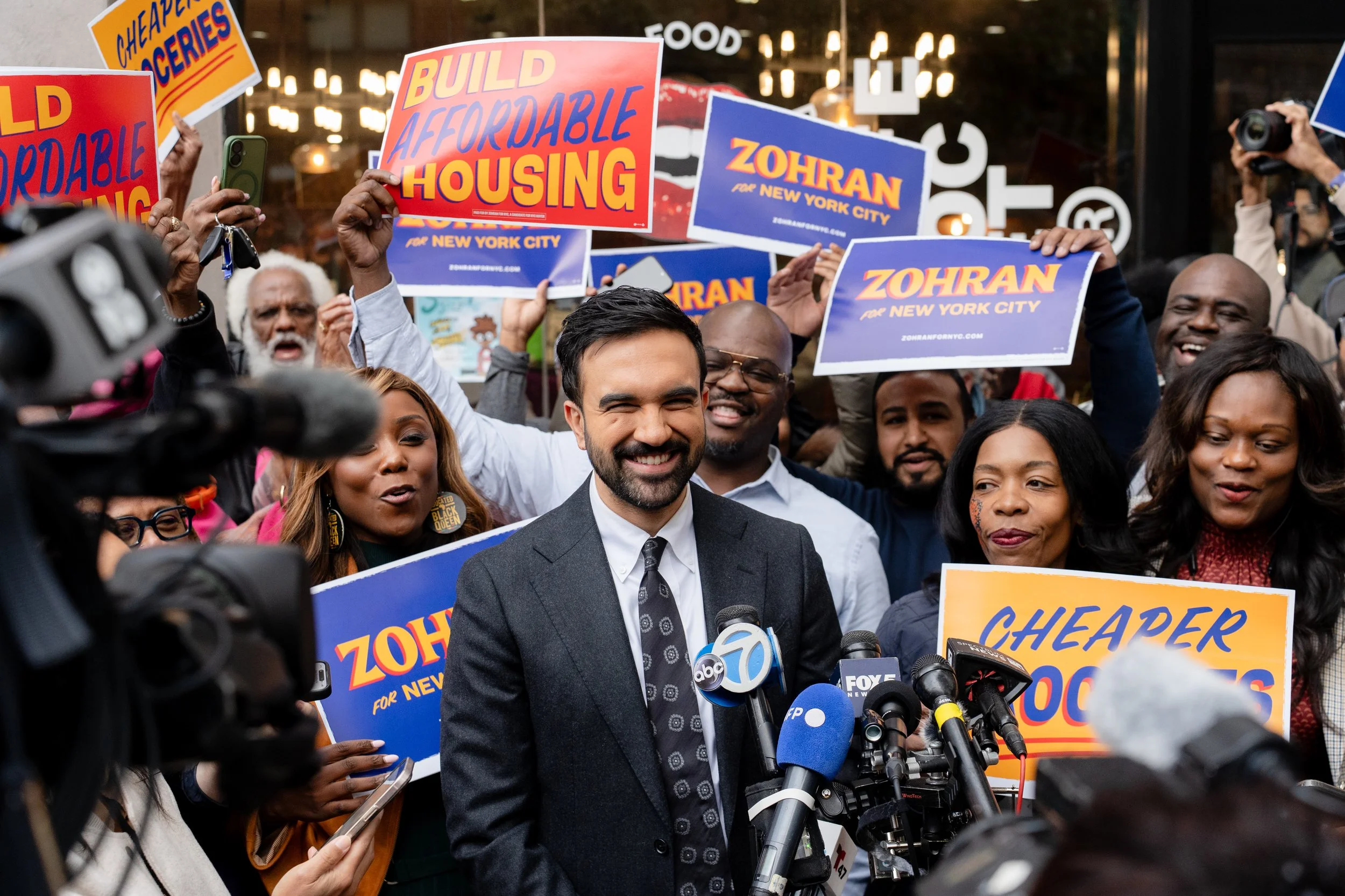 A group of diverse people protesting for affordable housing, holding signs that say 'Build Affordable Housing,' 'Cheaper Grocery,' and 'Zohar for New York City,' with a man in a suit speaking at a microphone in front of the crowd.