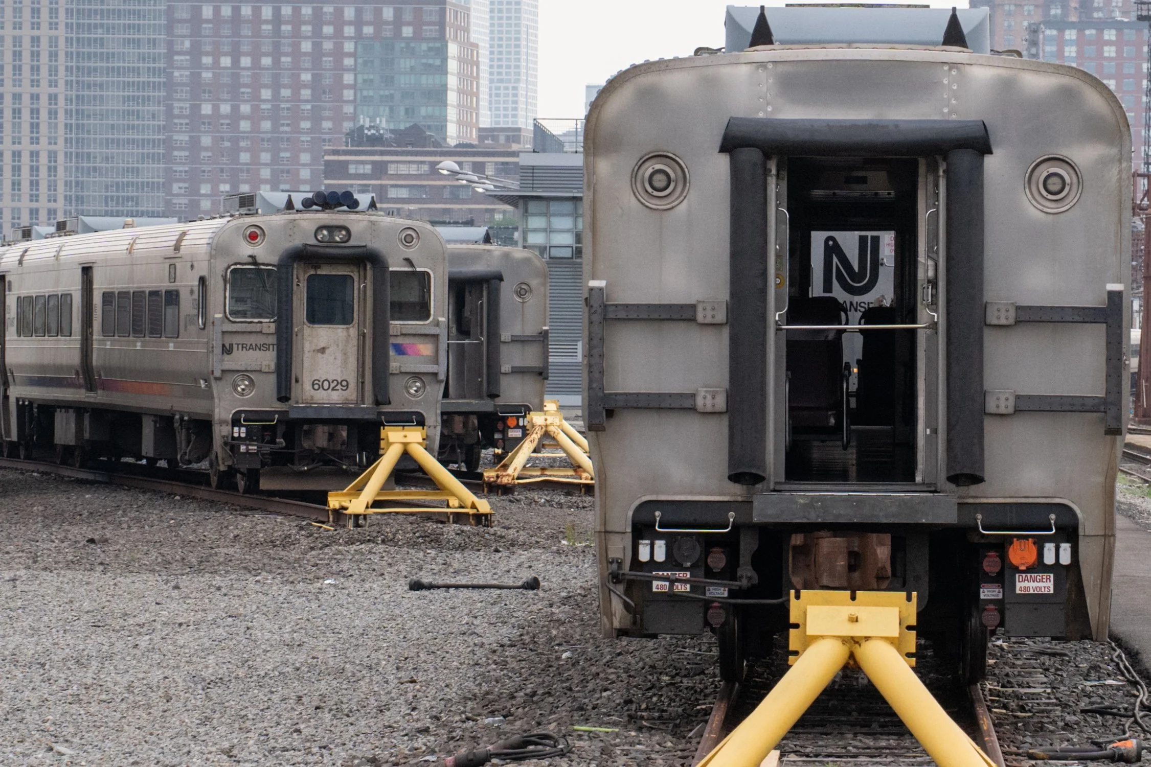 Two silver NJ Transit trains on parallel tracks in an urban train yard with tall buildings in the background. One train is front-facing, and the rear of the other is visible from the back, with yellow wheel chocks and safety signs nearby.
