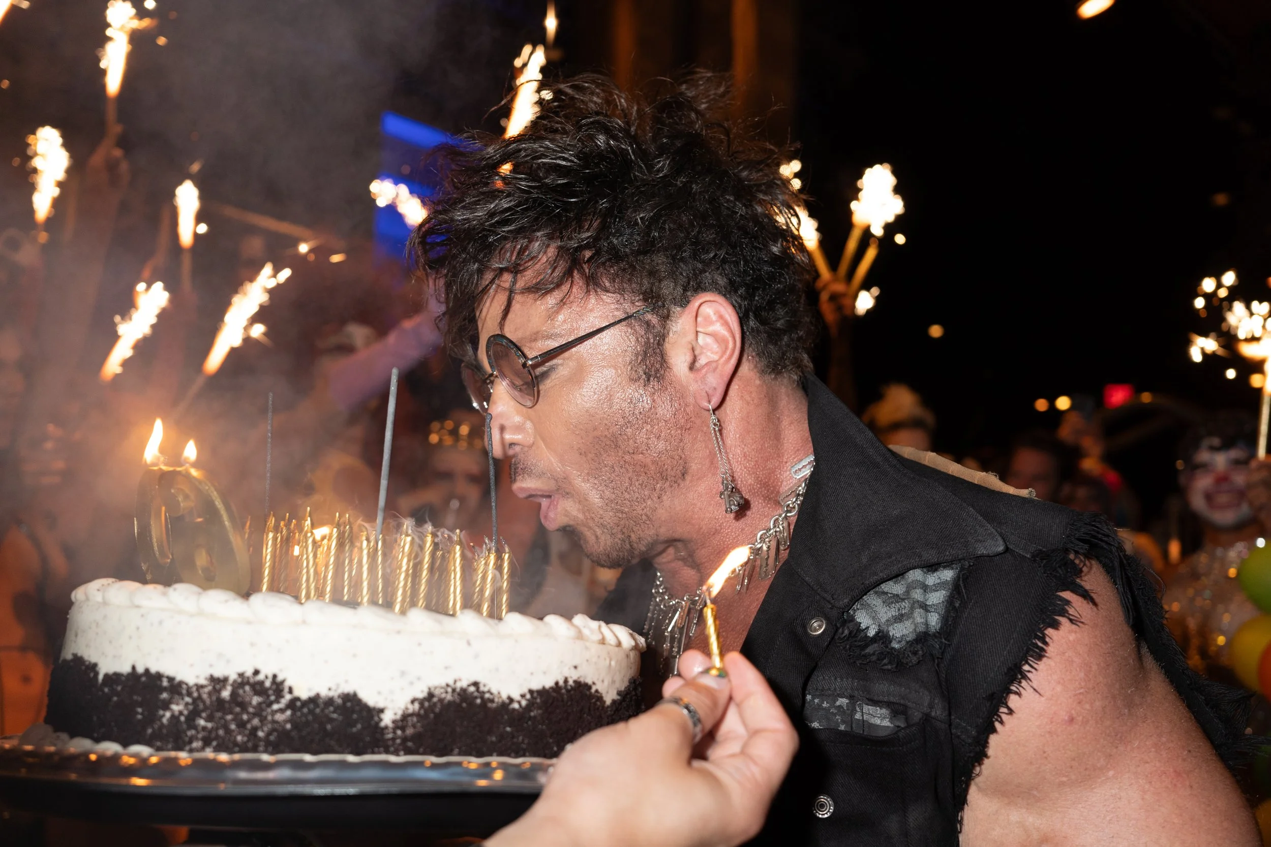 A man with curly hair, wearing sunglasses and earrings, is blowing out candles on a birthday cake during a celebration at night. There are sparklers and people in the background, some with face paint and costumes.