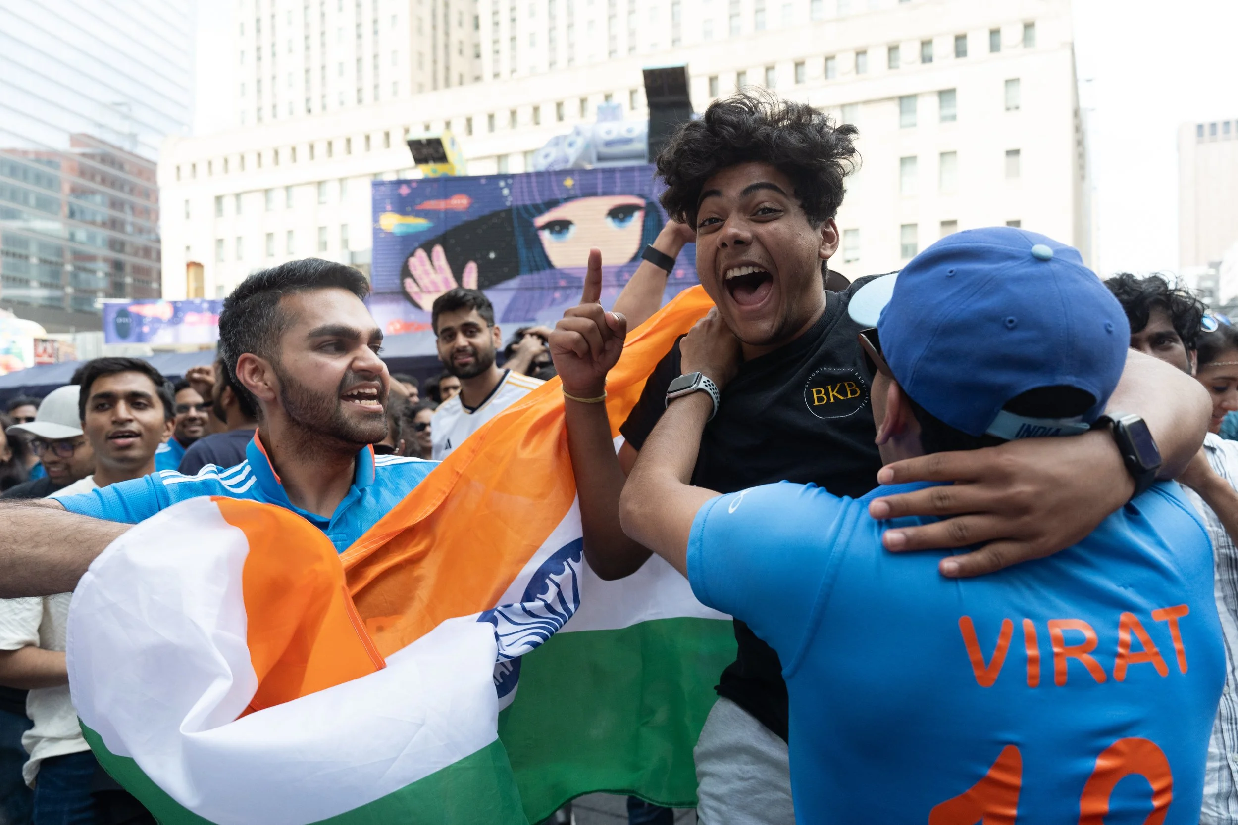 A group of jubilant Indian cricket fans celebrating at an outdoor event, with some holding the Indian flag, in a city setting with tall buildings and digital screens in the background.