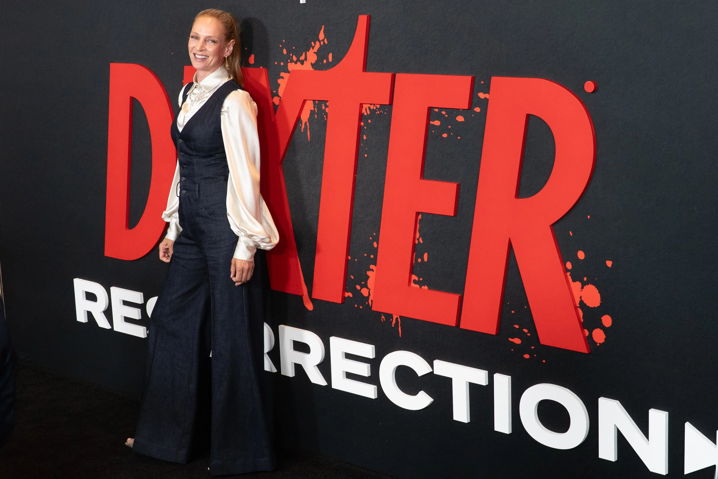 A woman with long red hair smiling, wearing a cream-colored satin blouse with puffed sleeves and a black vest, standing in front of a "DARK TOWER" promotional backdrop.