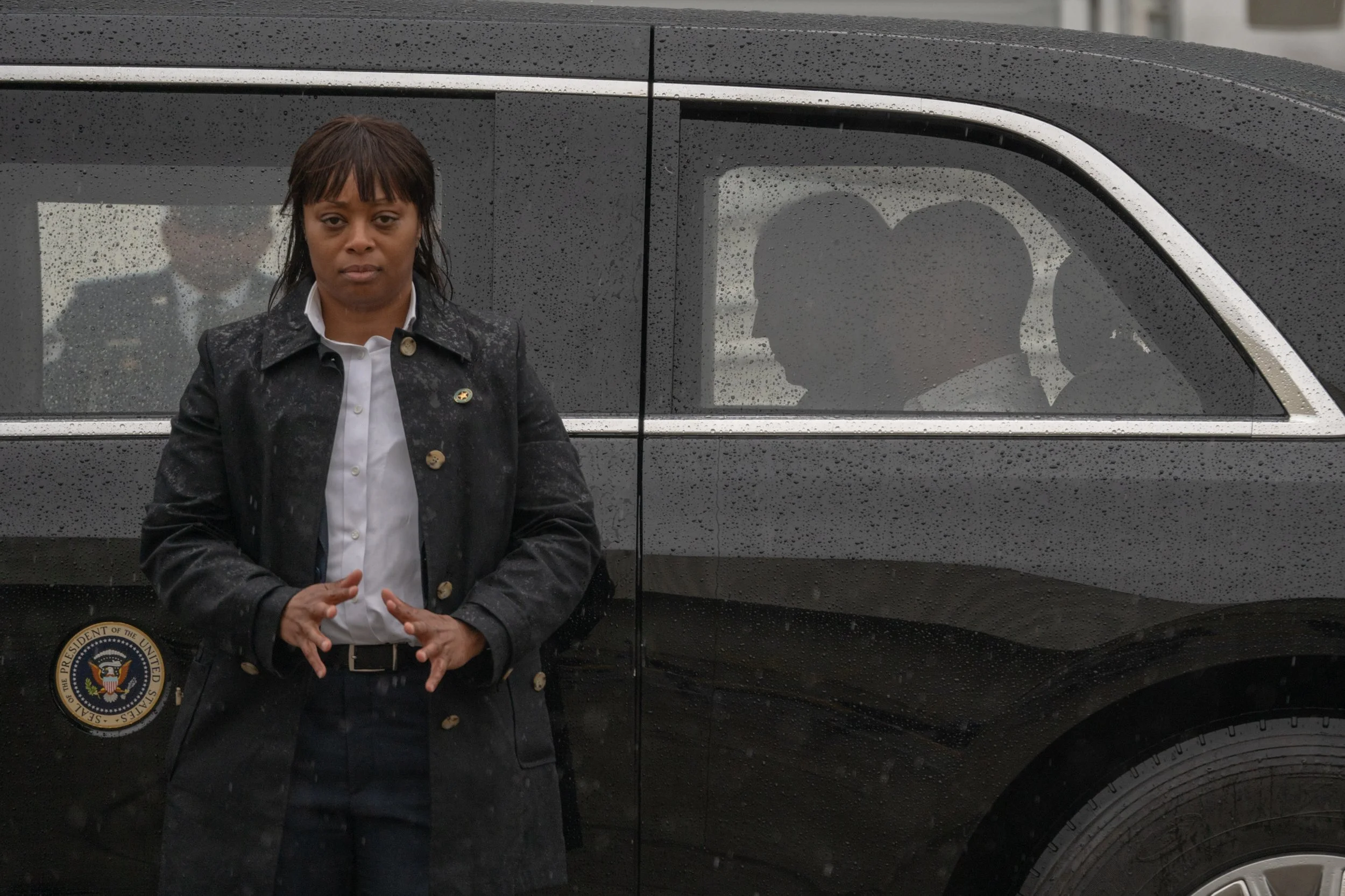 A woman with dark hair and a black coat stands in front of a dark vehicle with rain droplets on it. Inside the vehicle, two people are seated and visible through the window, facing each other and talking.