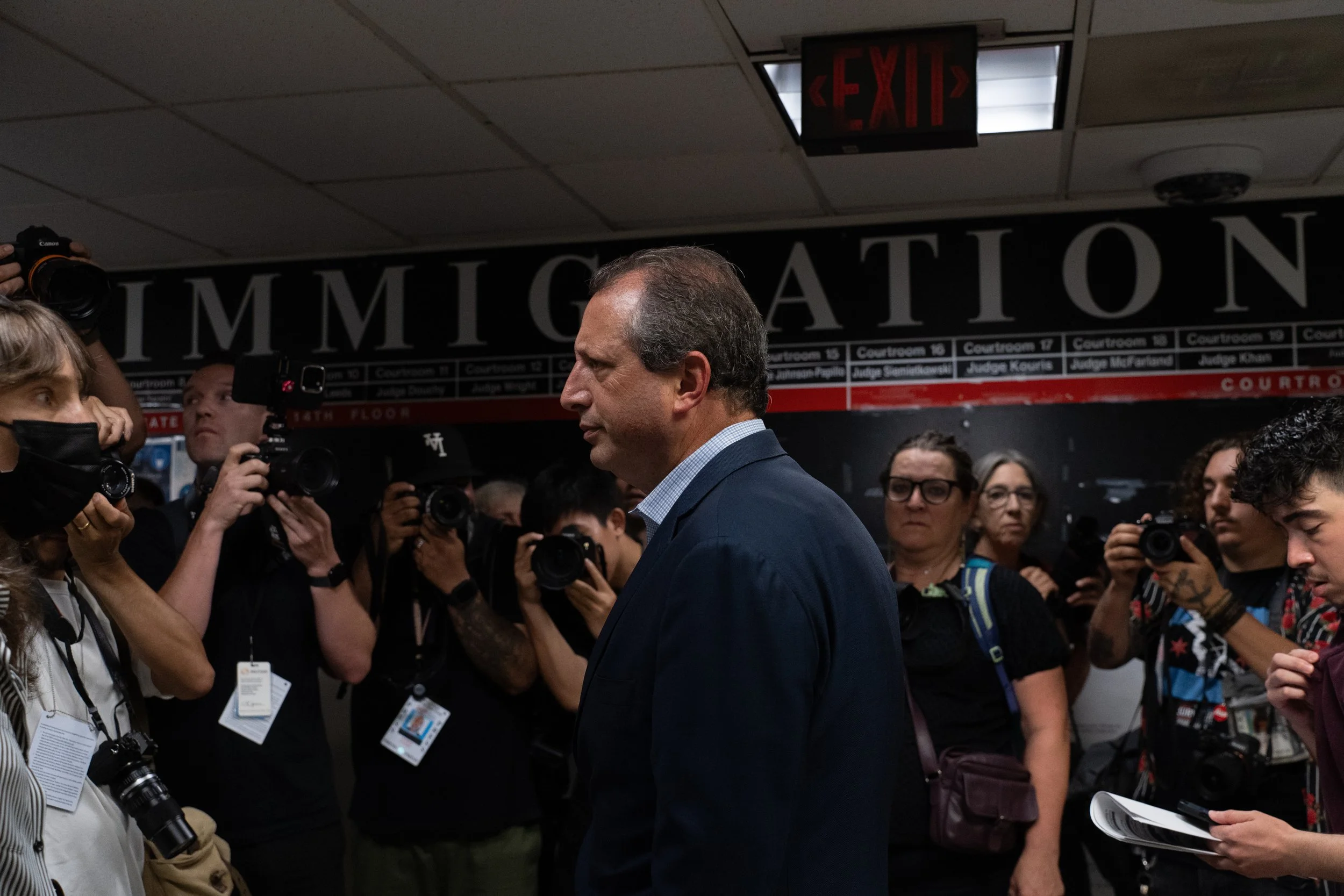 A man in a navy suit walking past a group of journalists and photographers in an indoor courthouse lobby, with a large sign showing courtrooms behind him.