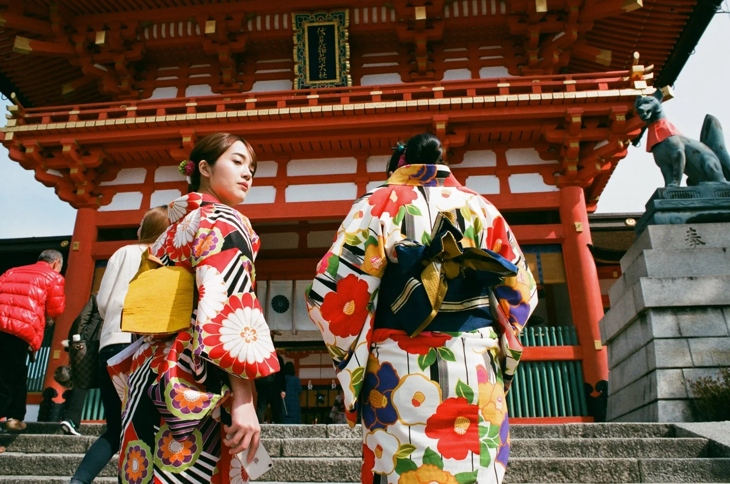 Two women in colorful kimonos standing on stone steps in front of a traditional Japanese shrine with red and white architecture, with other visitors around.