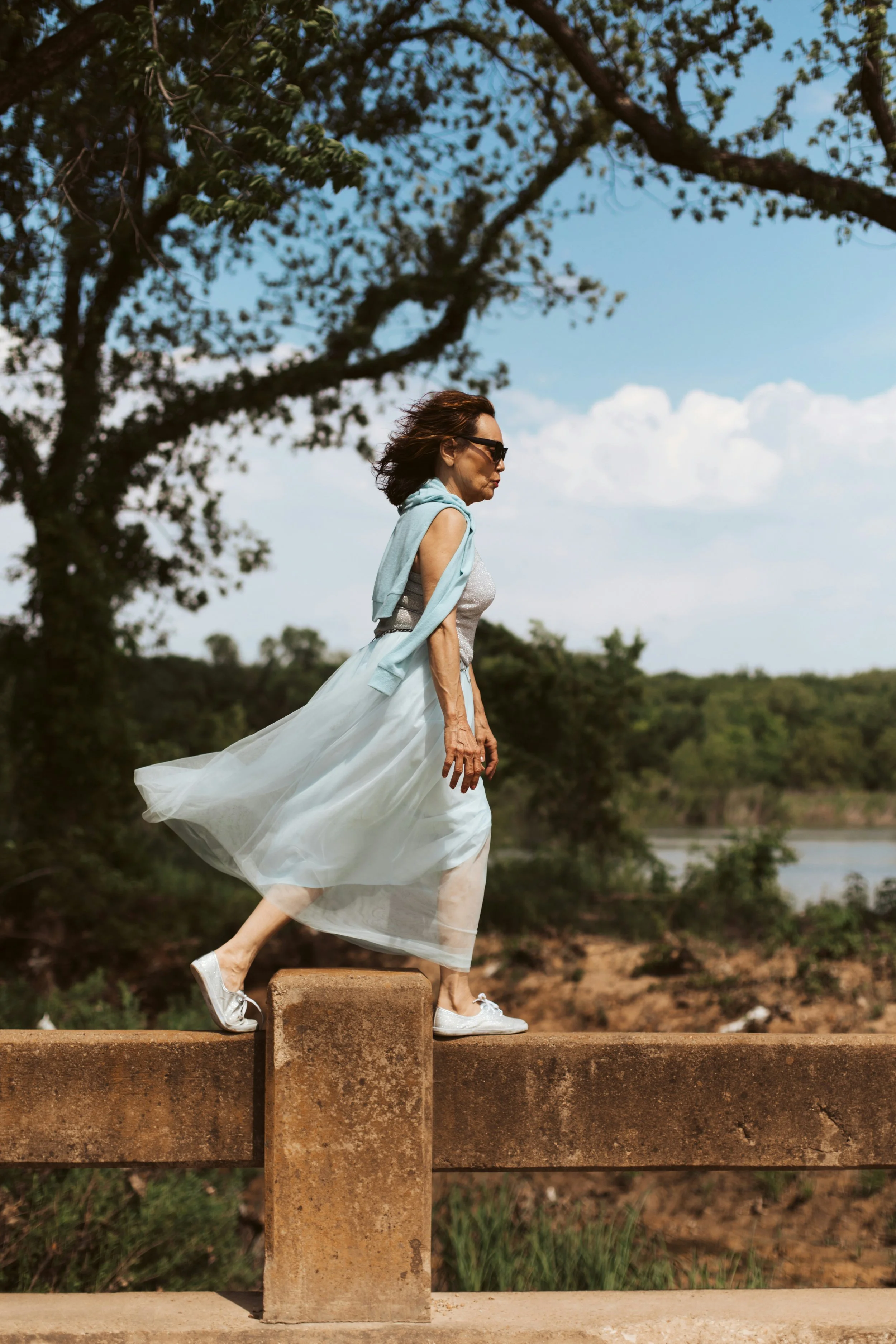 A woman wearing a white dress, gray top, and sneakers walking along a concrete barrier outdoors with trees and a blue sky in the background.