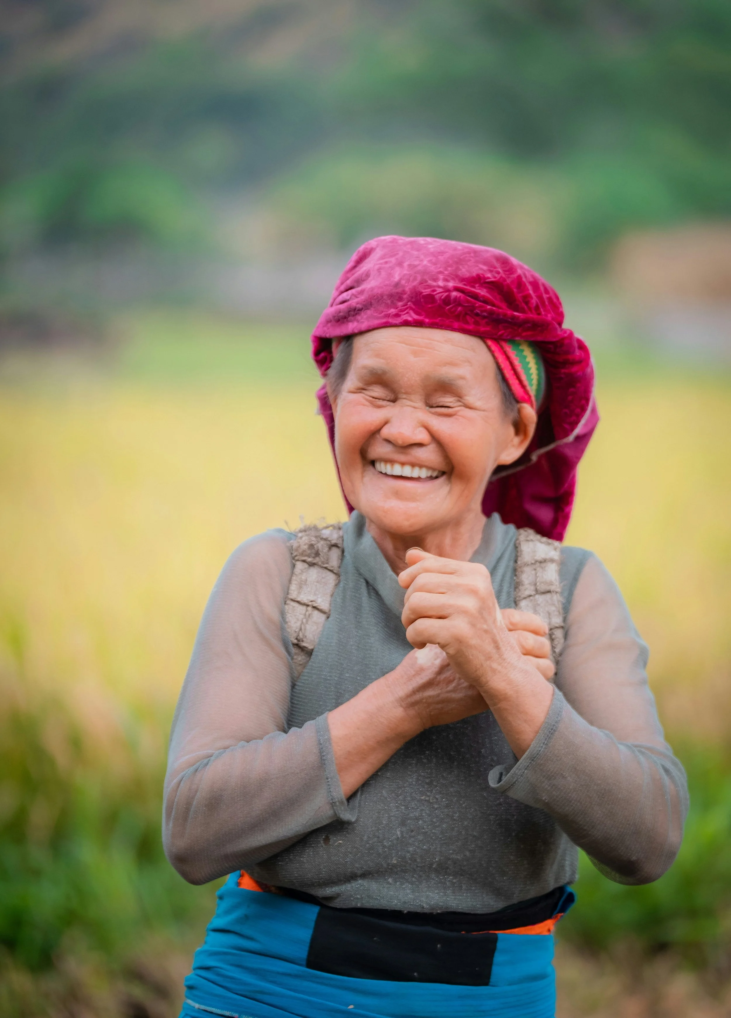 An elderly woman with a big smile, wearing a bright pink headscarf and gray hiking clothes, stands outdoors in a green field.