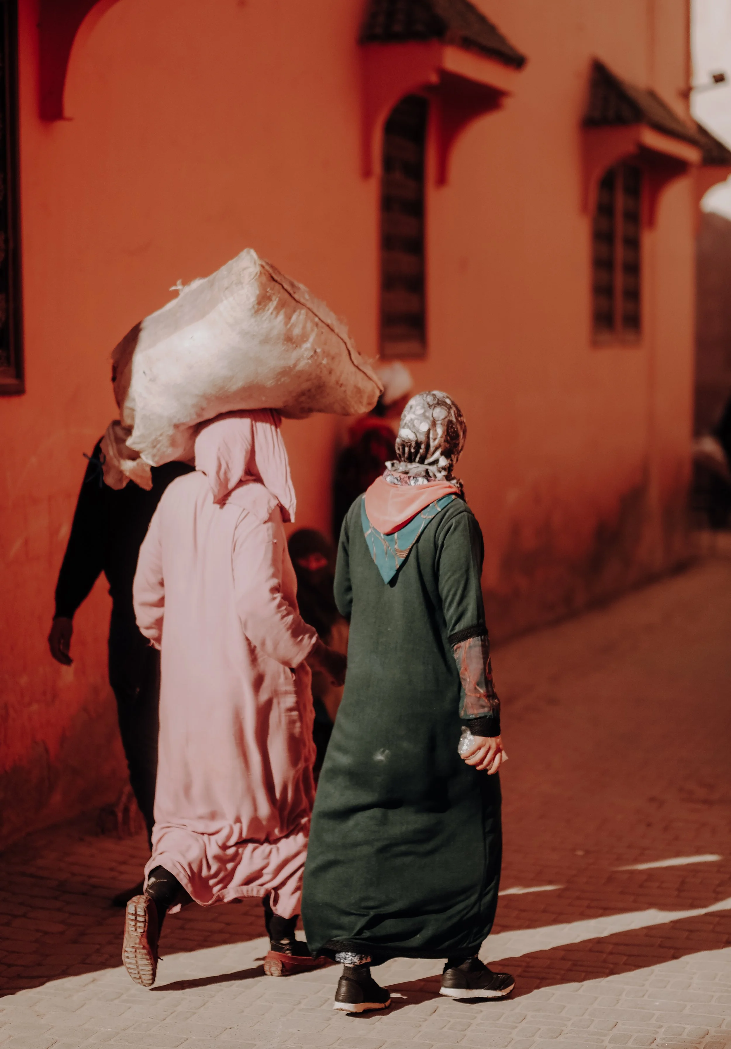 Two women walking on a cobblestone street at night, one carrying a large load on her head, with a peach-colored building in the background.