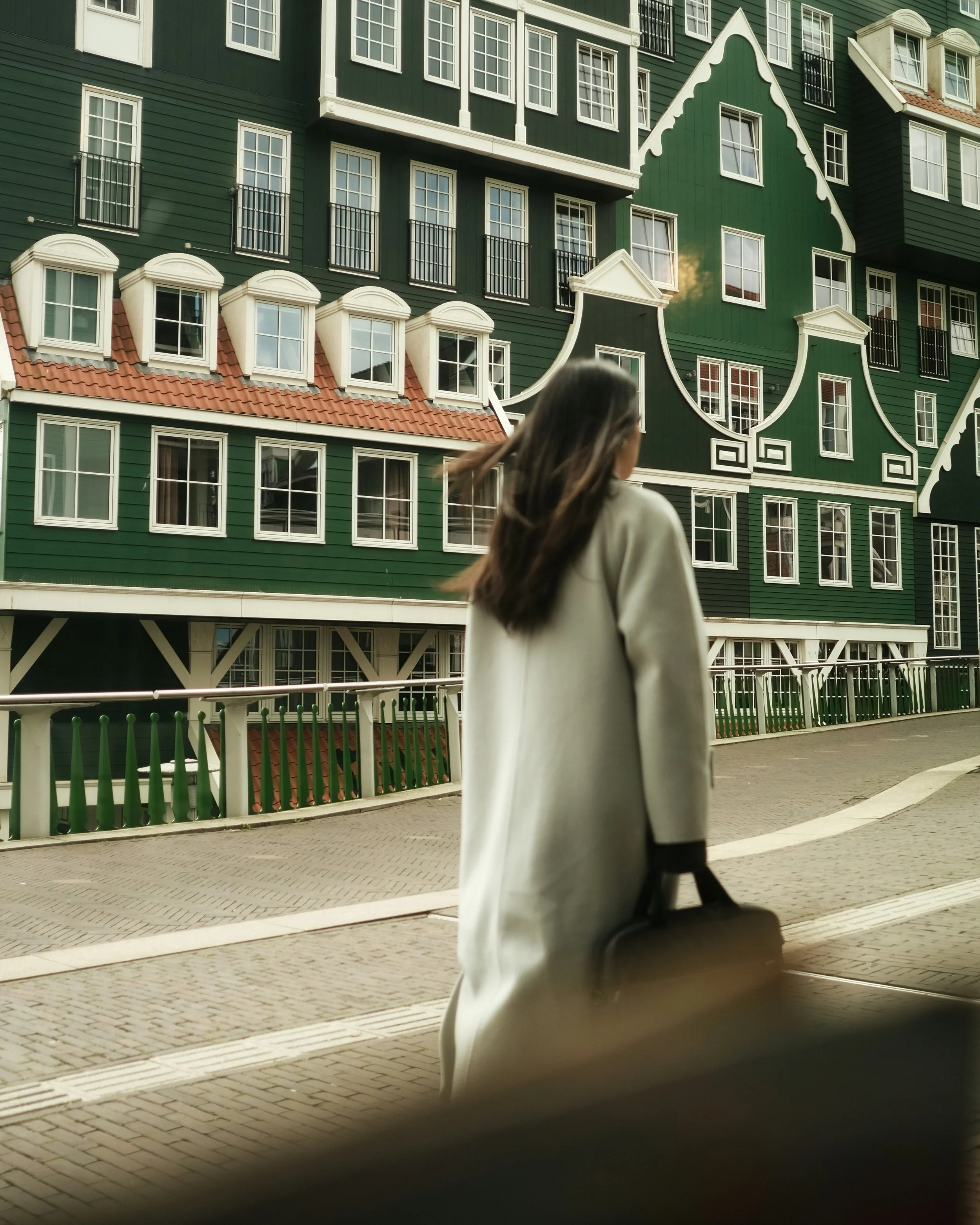 A woman with long dark hair, wearing a white coat, carrying a black bag, walking along a paved sidewalk in front of a large green building with white-trimmed windows and decorative architecture.
