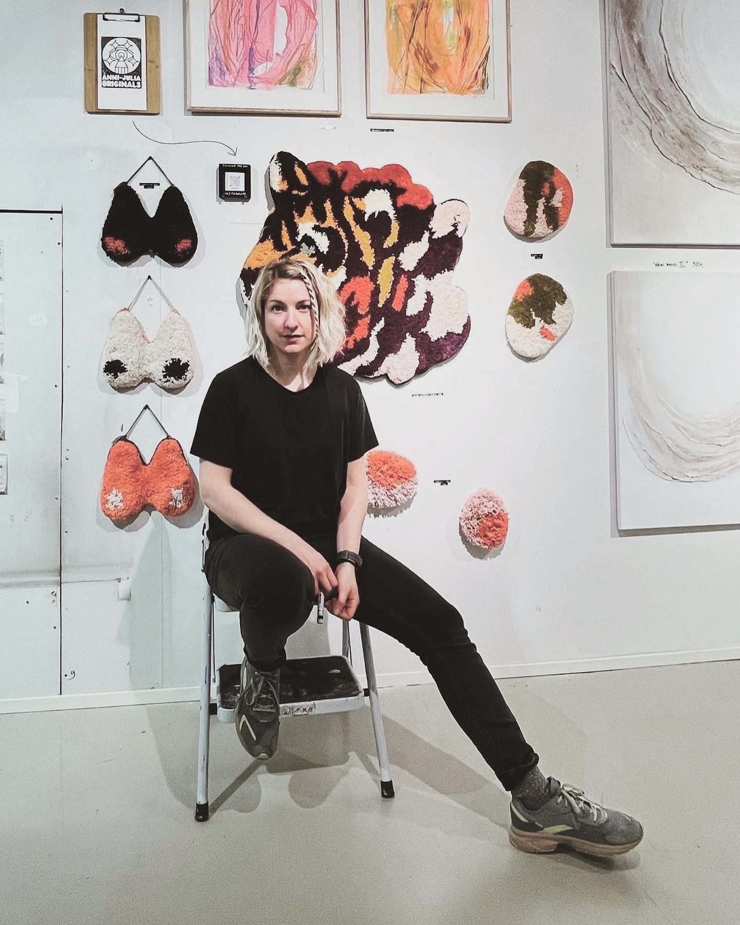 Nuori nainen istuu tuolilla taiteenäyttelyn edessä, taustalla on seinä täynnä värikkäitä teoksia ja tekstiä.