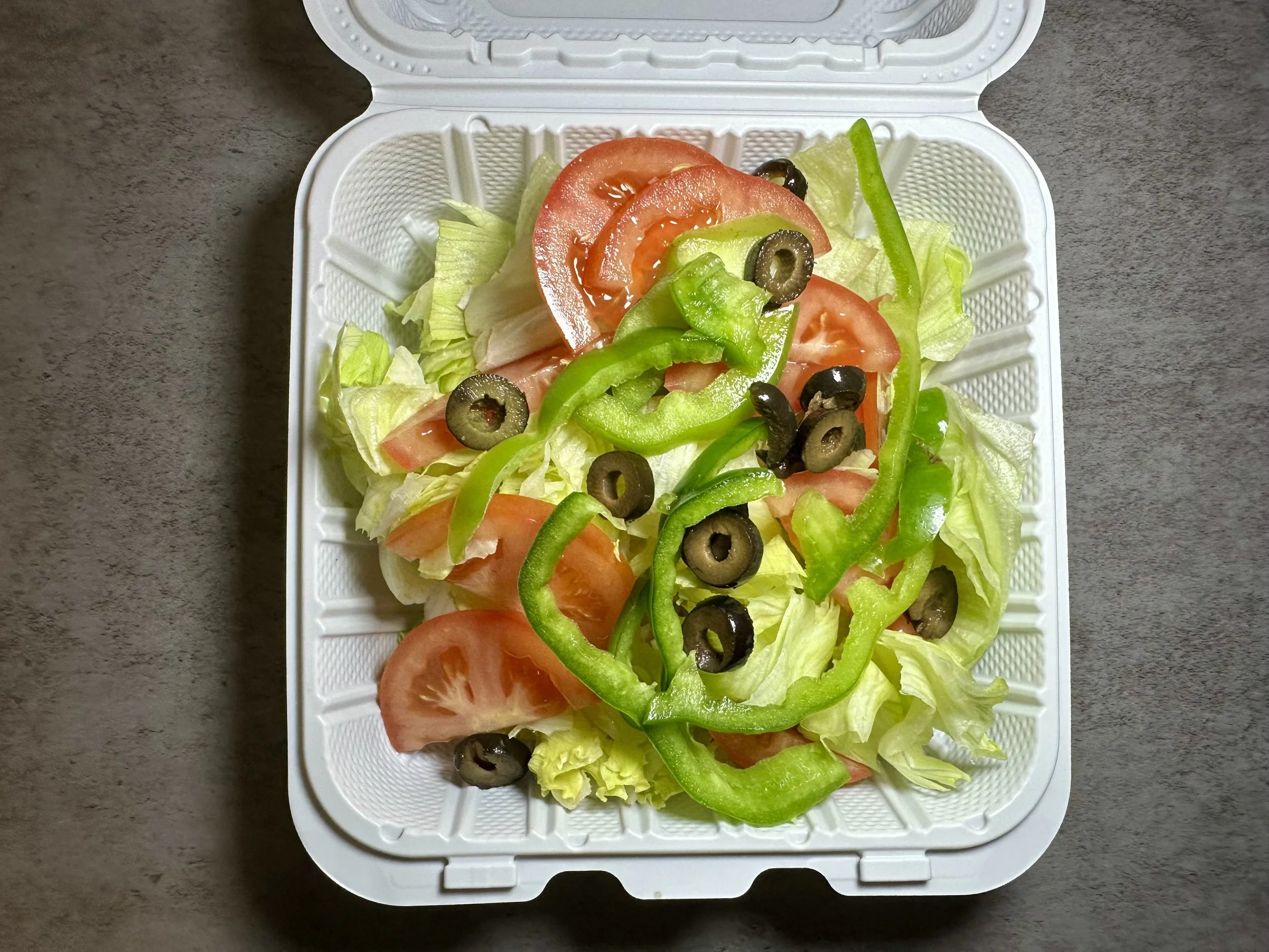 A salad with sliced tomatoes, chopped lettuce, sliced green bell peppers, and black olives in a white disposable container.