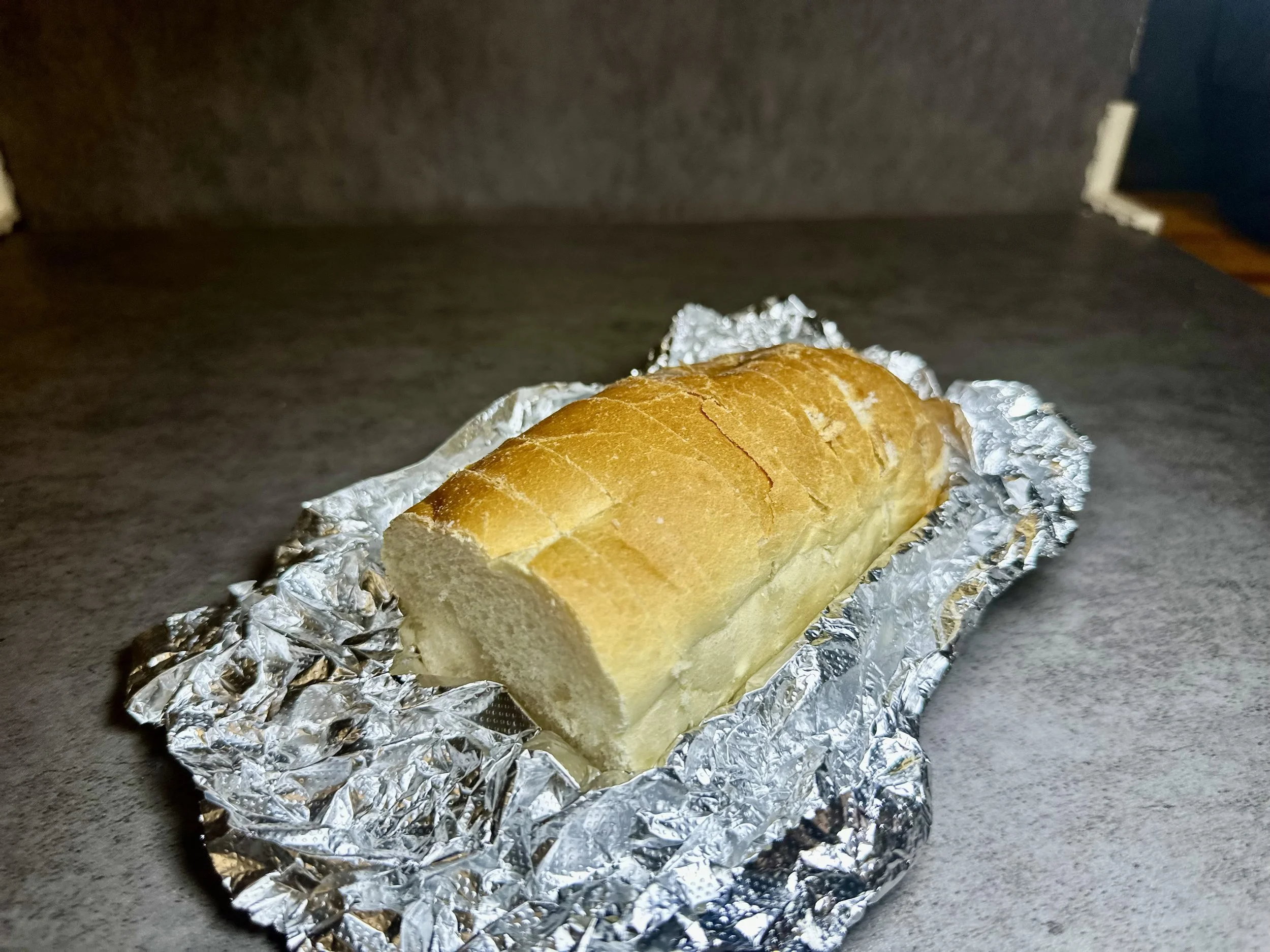 A loaf of bread partially sliced, resting on crumpled aluminum foil on a dark countertop.