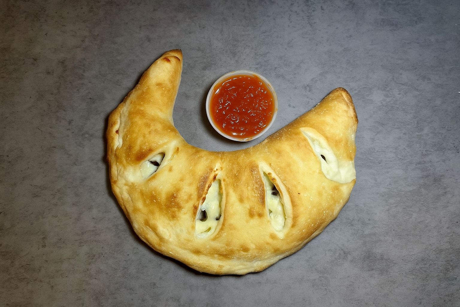 A bread crescent with cheese and jalapeño slices, served with a side of red sauce in a small container, on a gray surface.