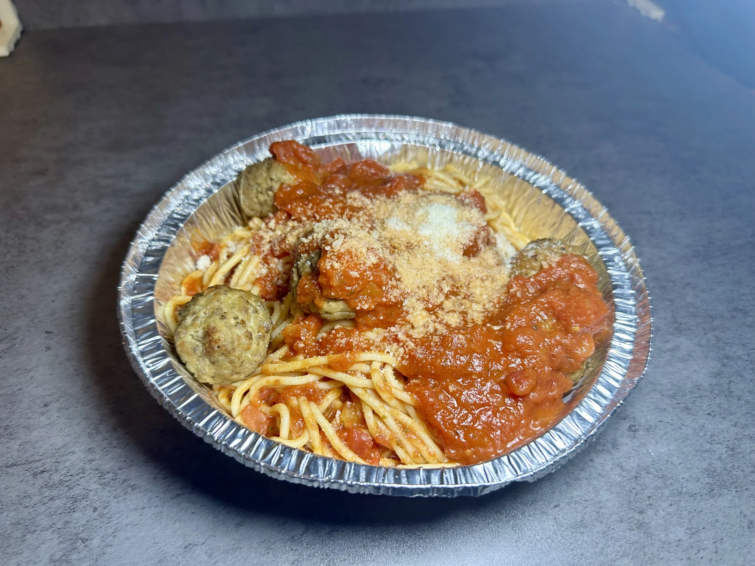 Foil takeout container with spaghetti, meatballs, marinara sauce, grated cheese, and seasonings on a dark surface.