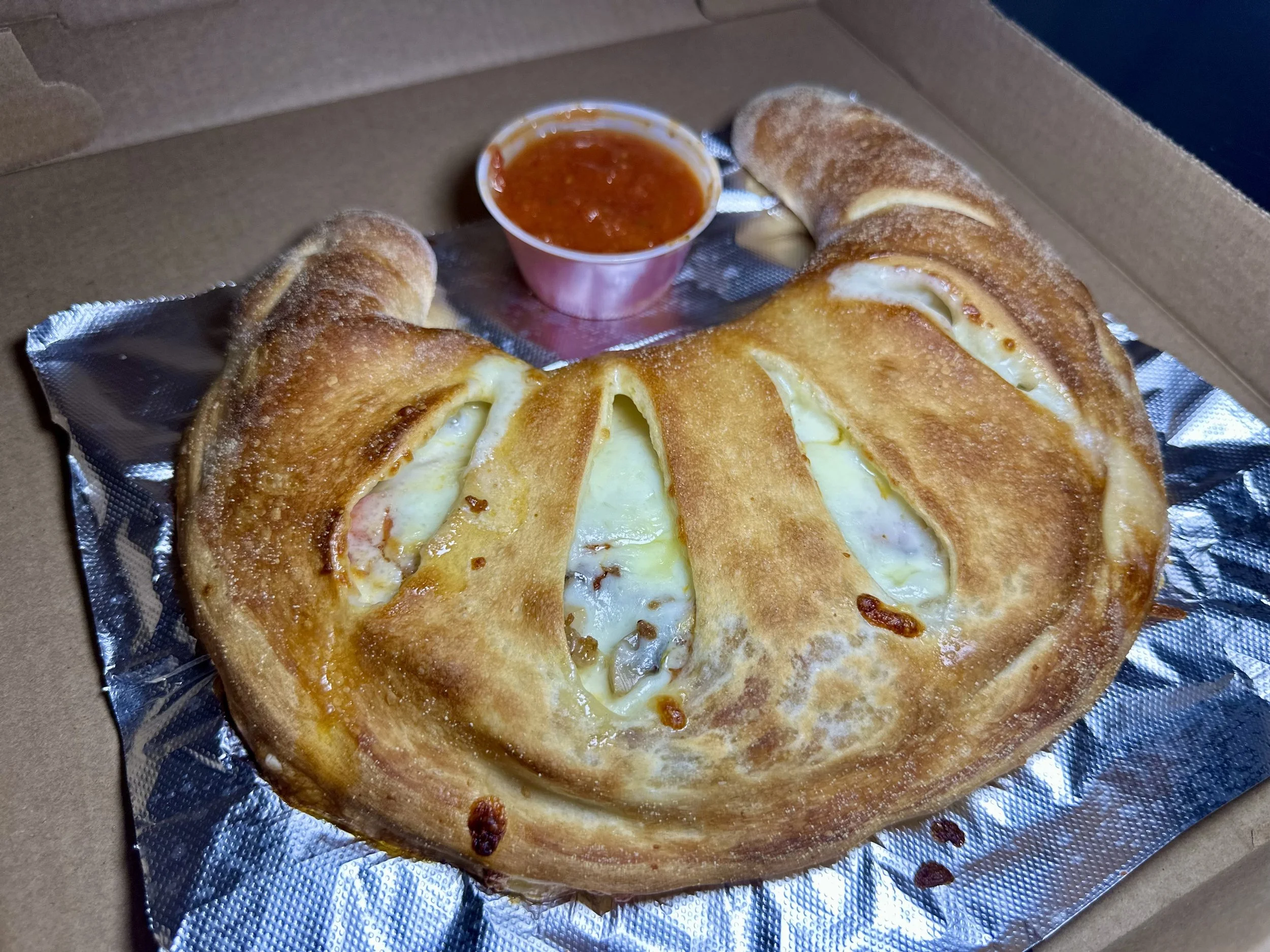 Baked calzone with melted cheese and crust, served in a cardboard box with a small cup of marinara sauce.