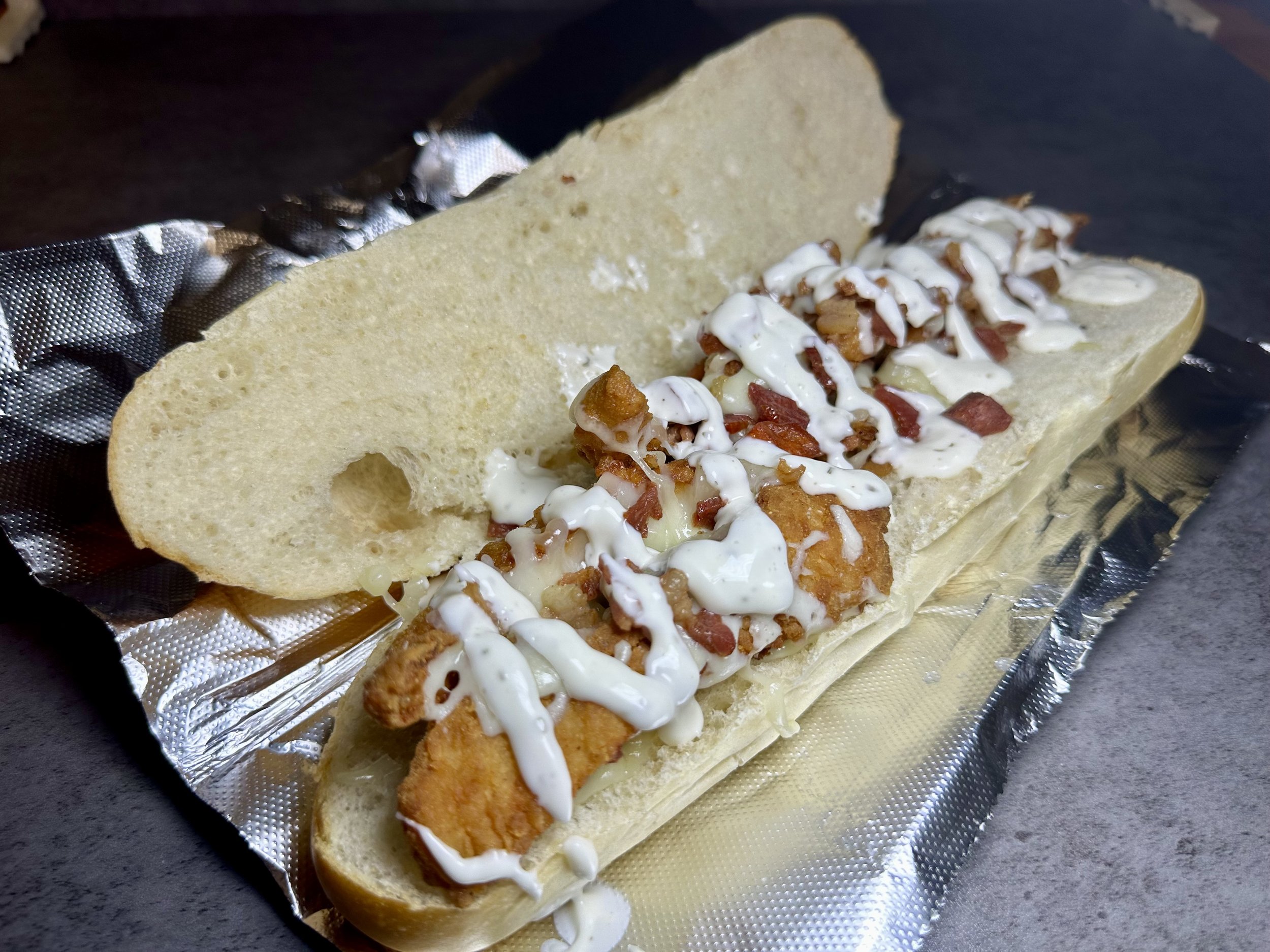 A fish sandwich in a hot dog bun with tartar sauce, served on foil