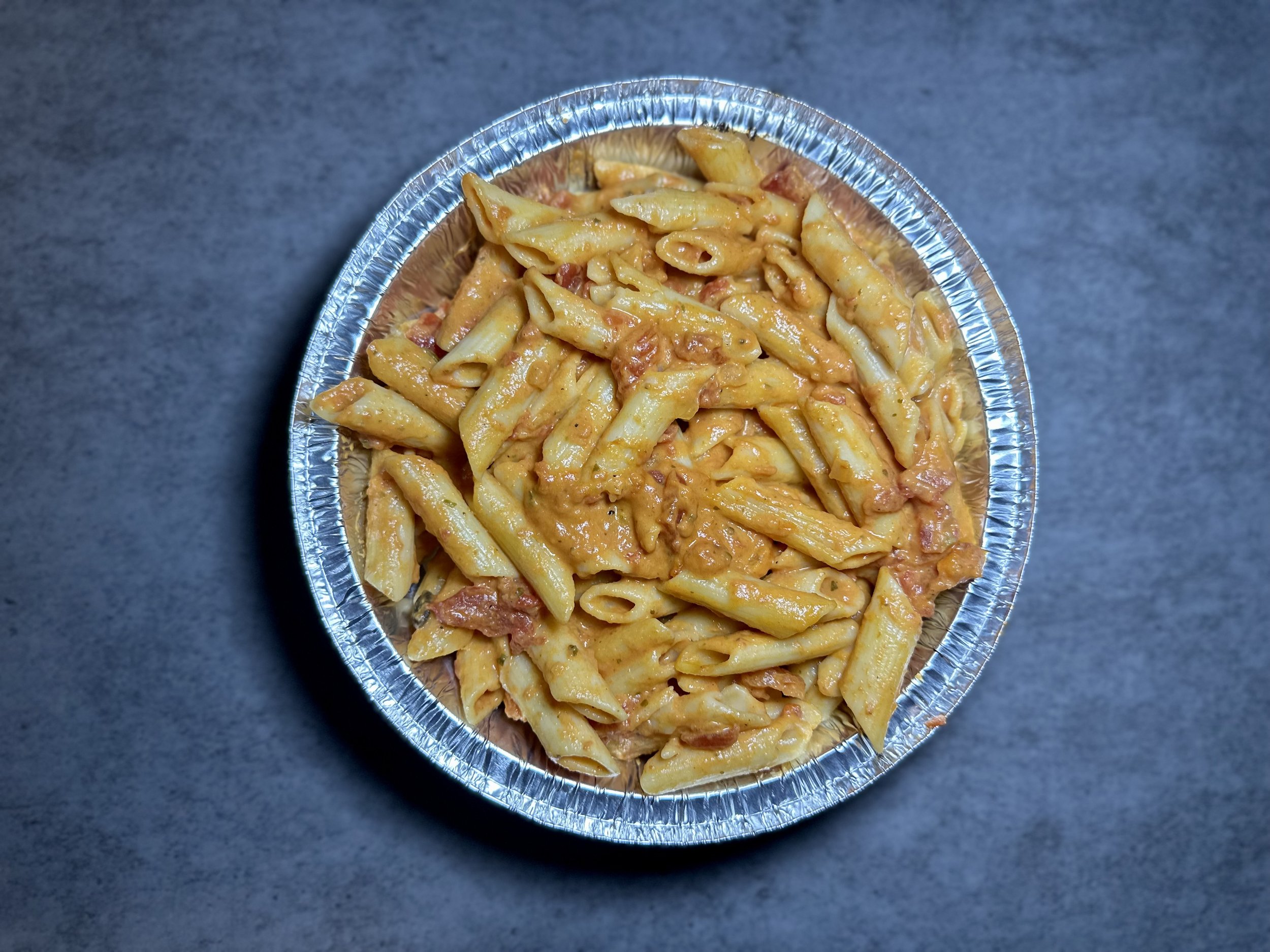 Disposable aluminum tray filled with cooked pasta with cheese and bacon bits on a dark gray surface.