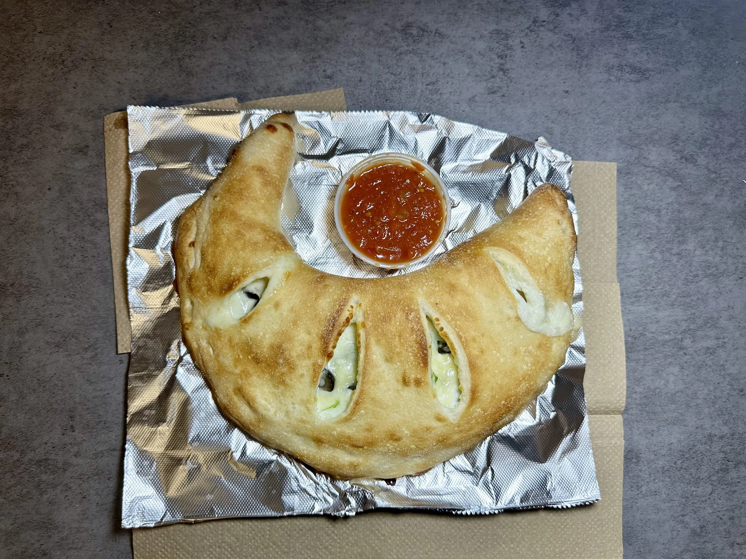 Deep-fried calzone with cheese and vegetables, served with a side of red marinara sauce in a small container, on crumpled foil on a textured gray surface.