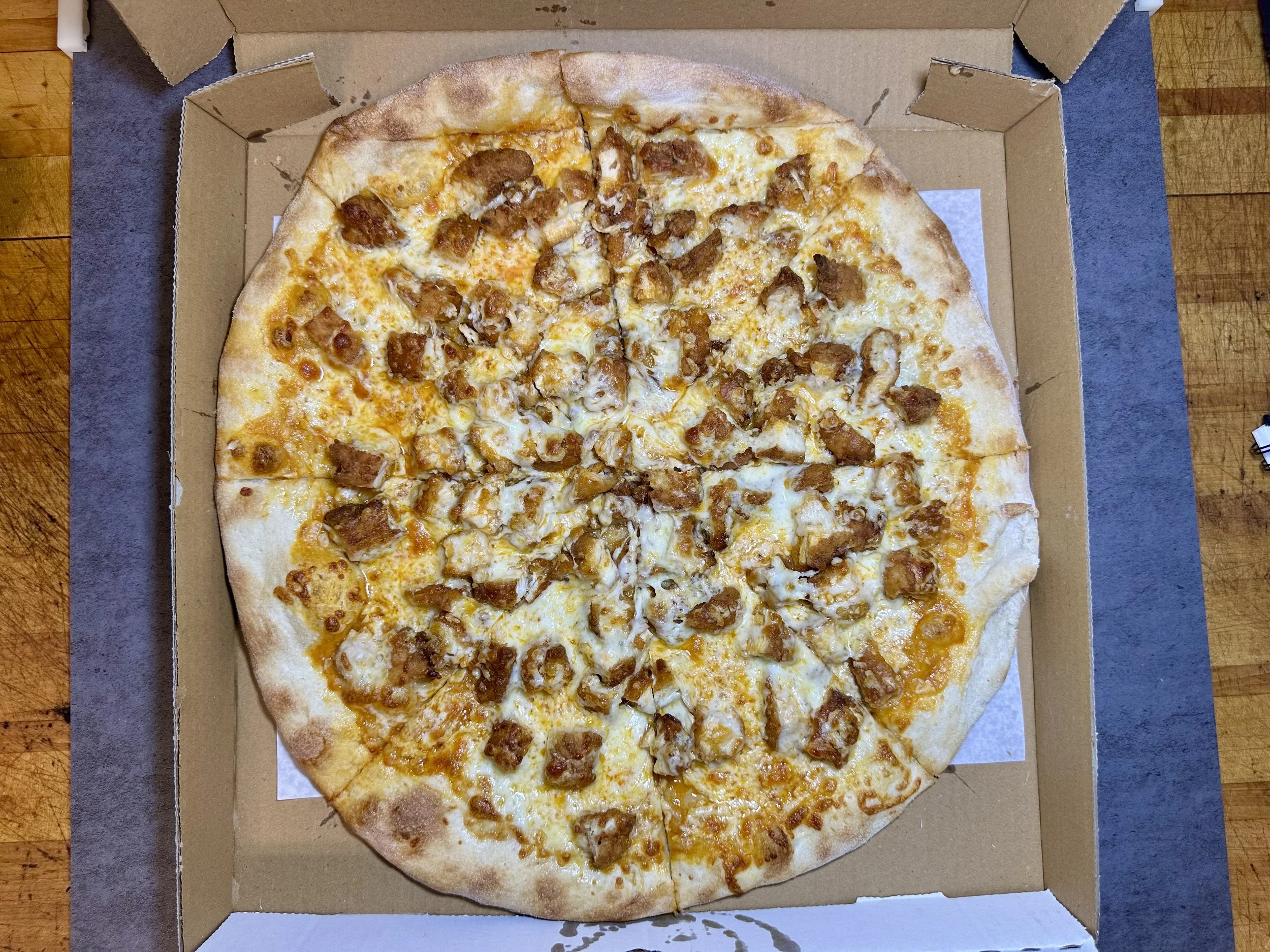 A cheese pizza with chicken toppings in a pizza box on a wooden table.