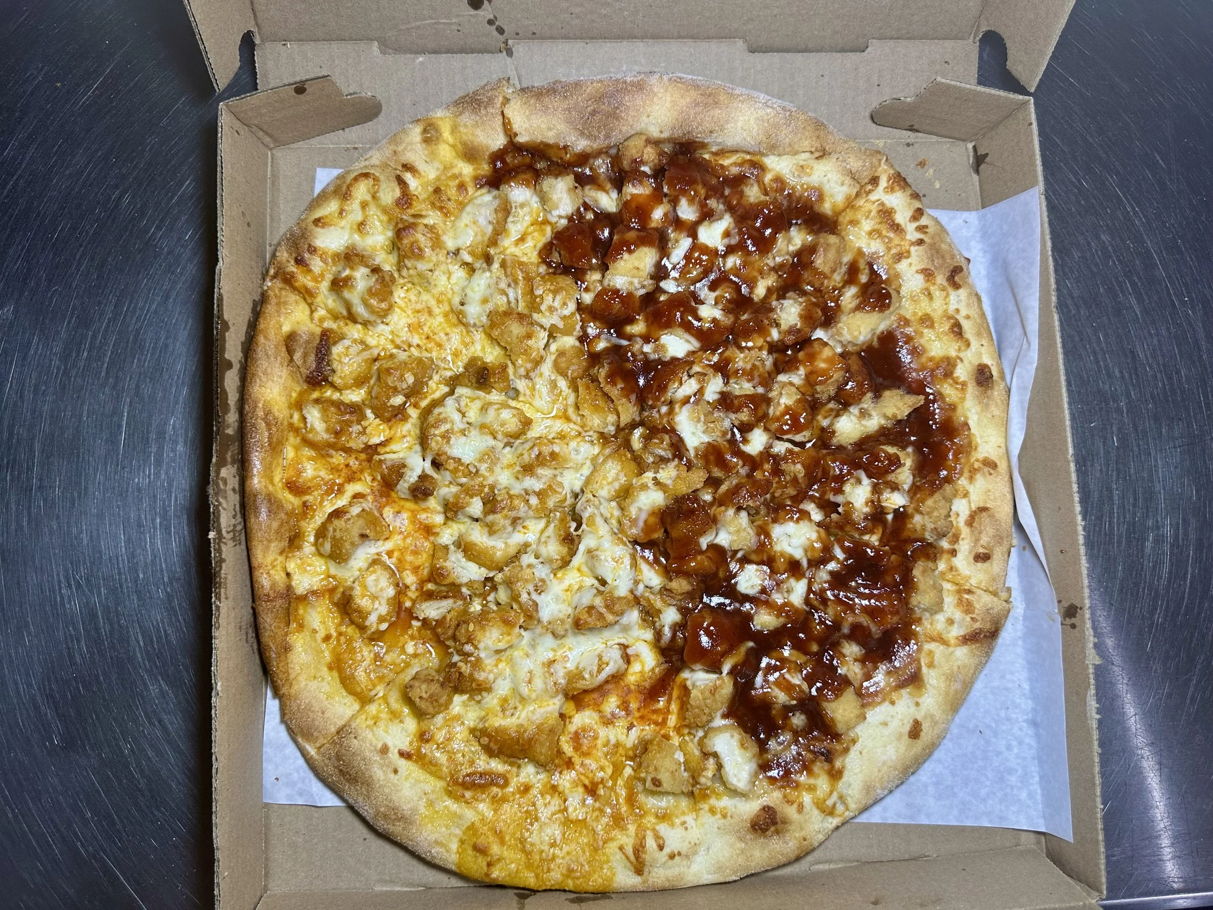 A pizza with half chicken and cheese and half barbecue chicken in a cardboard box on a metal surface.