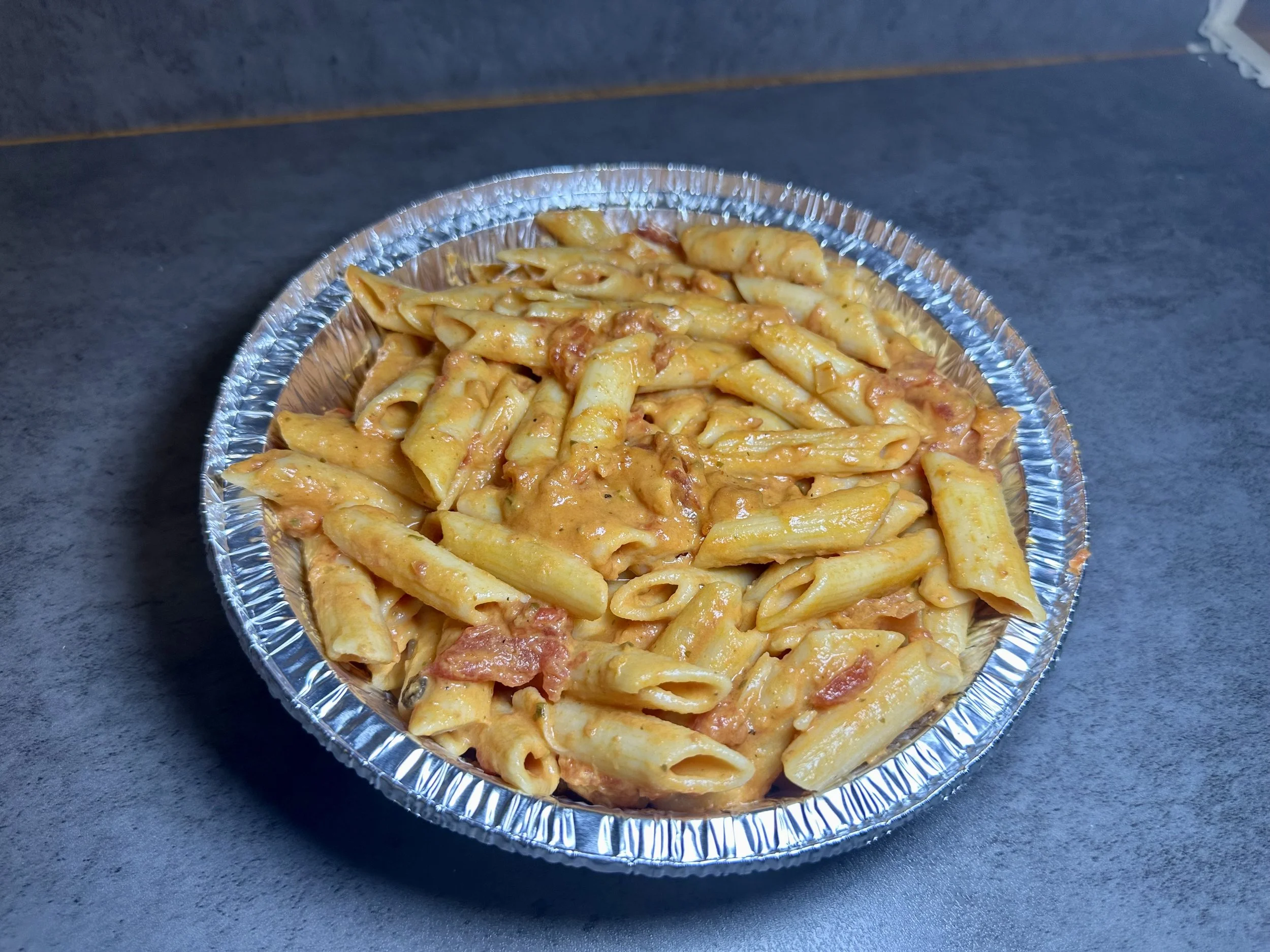 Foil container of baked penne pasta with tomato sauce and melted cheese on a dark surface.