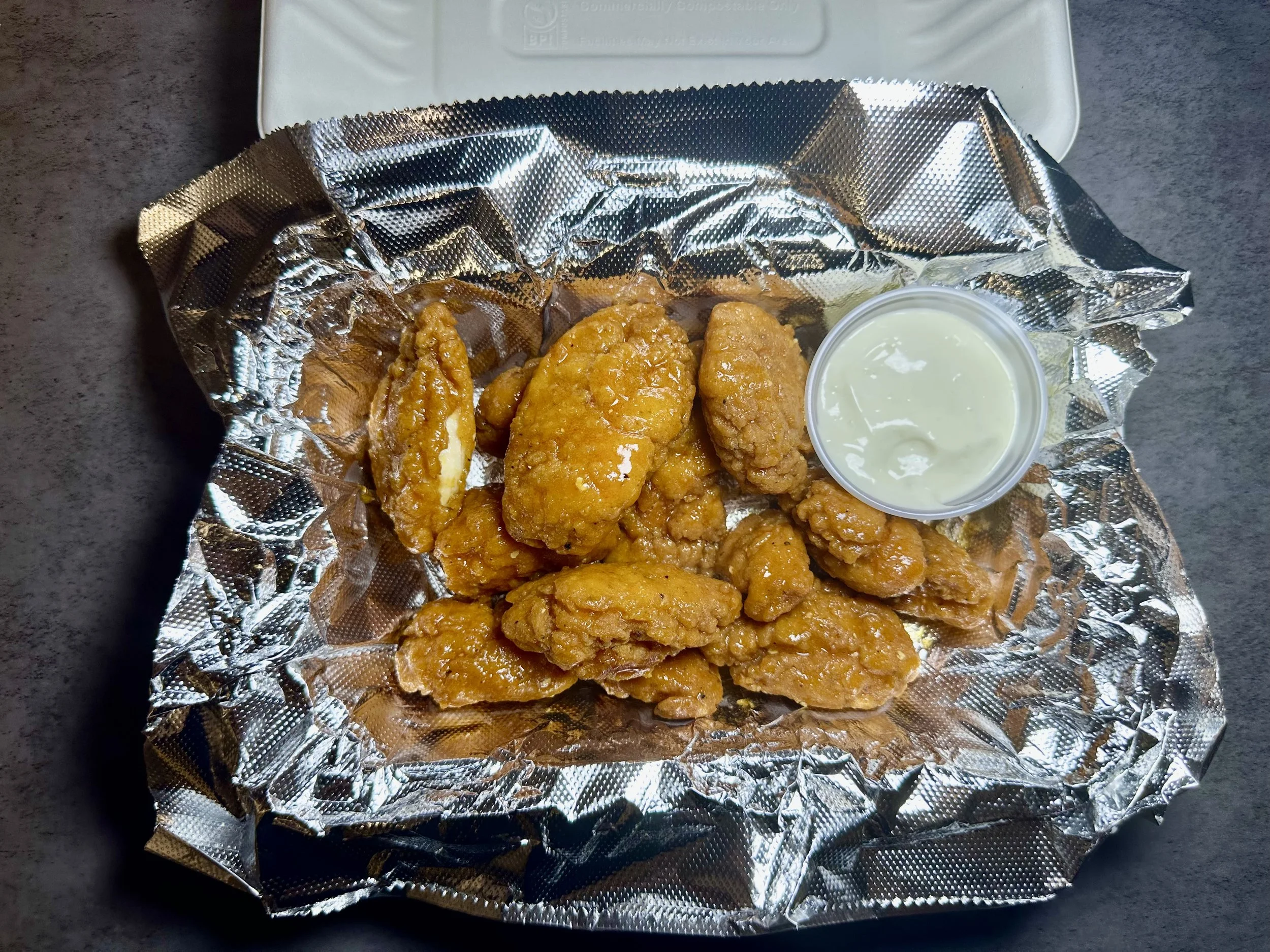 Fried chicken pieces in foil with a small cup of ranch dressing on a dark surface.