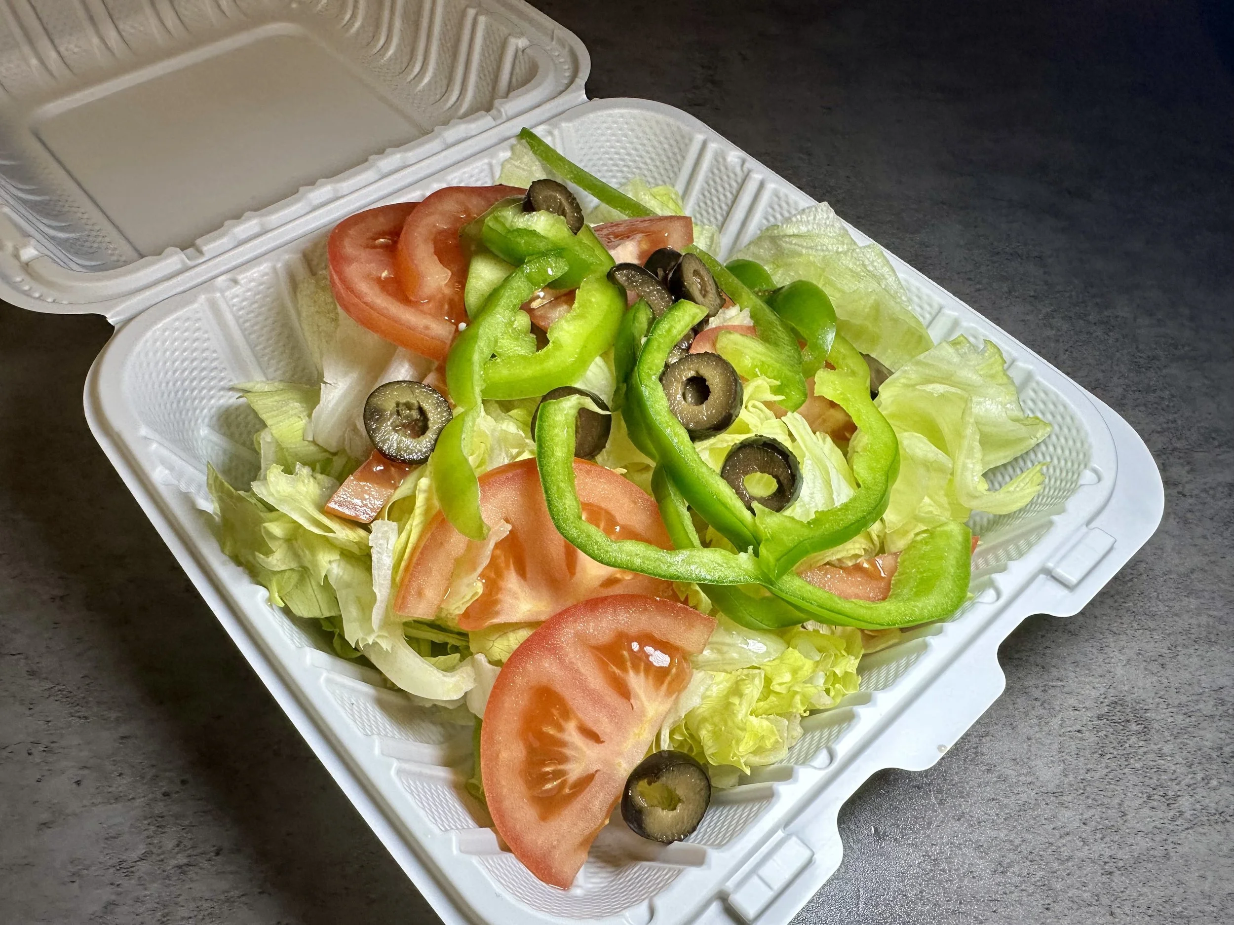 A styrofoam takeout container filled with a fresh vegetable salad including sliced tomatoes, chopped lettuce, sliced green bell peppers, and black olives, placed on a dark surface.