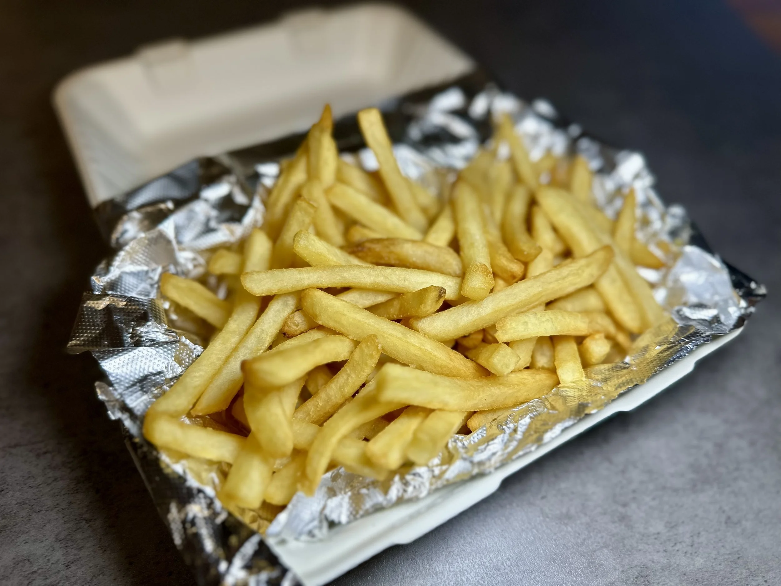 A small packet of French fries on a black surface, with the fries inside partially covered by aluminum foil.