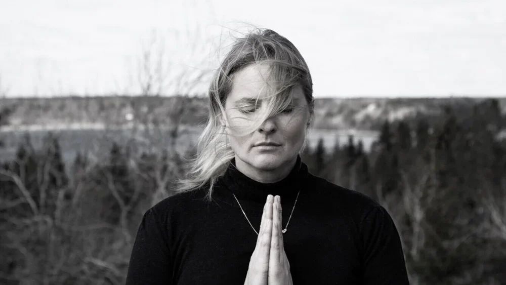 A woman with blonde hair, wearing a black turtleneck, stands outdoors with her hands pressed together in a prayer pose, eyes closed, in front of a natural landscape with trees and a cloudy sky.