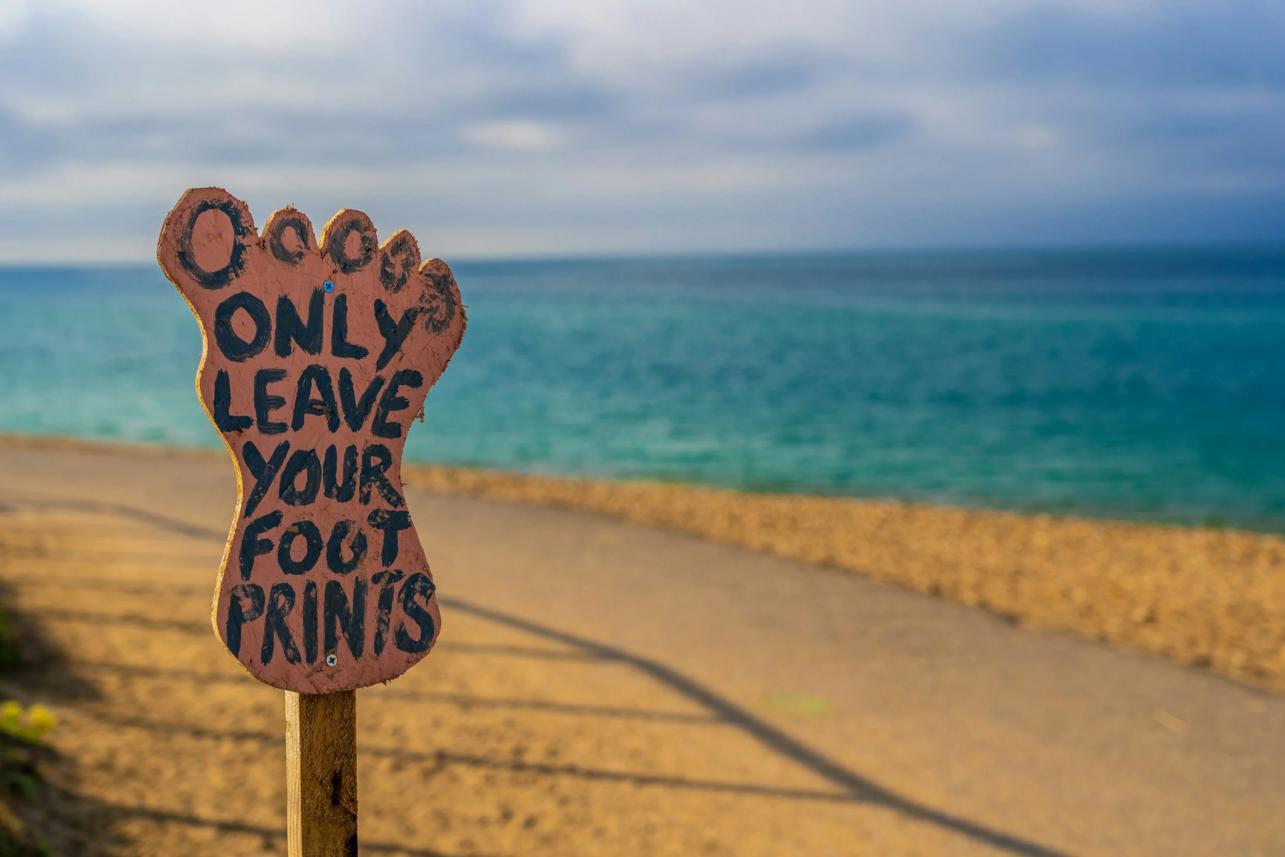 A sign shaped like a foot with the words 'Only leave your foot prints' written on it, situated on a sandy beach with ocean and cloudy sky in the background.