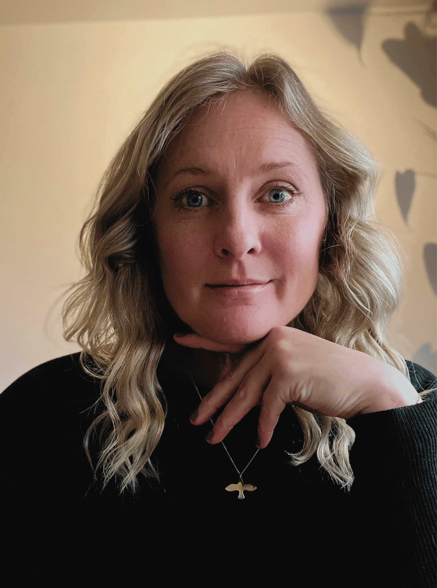 Portrait of a woman with blonde, wavy hair, blue eyes, wearing a black top and a gold necklace with a tree pendant, resting her chin on her hand, indoors with a light-colored wall and shadowed heart-shaped cutouts in the background.