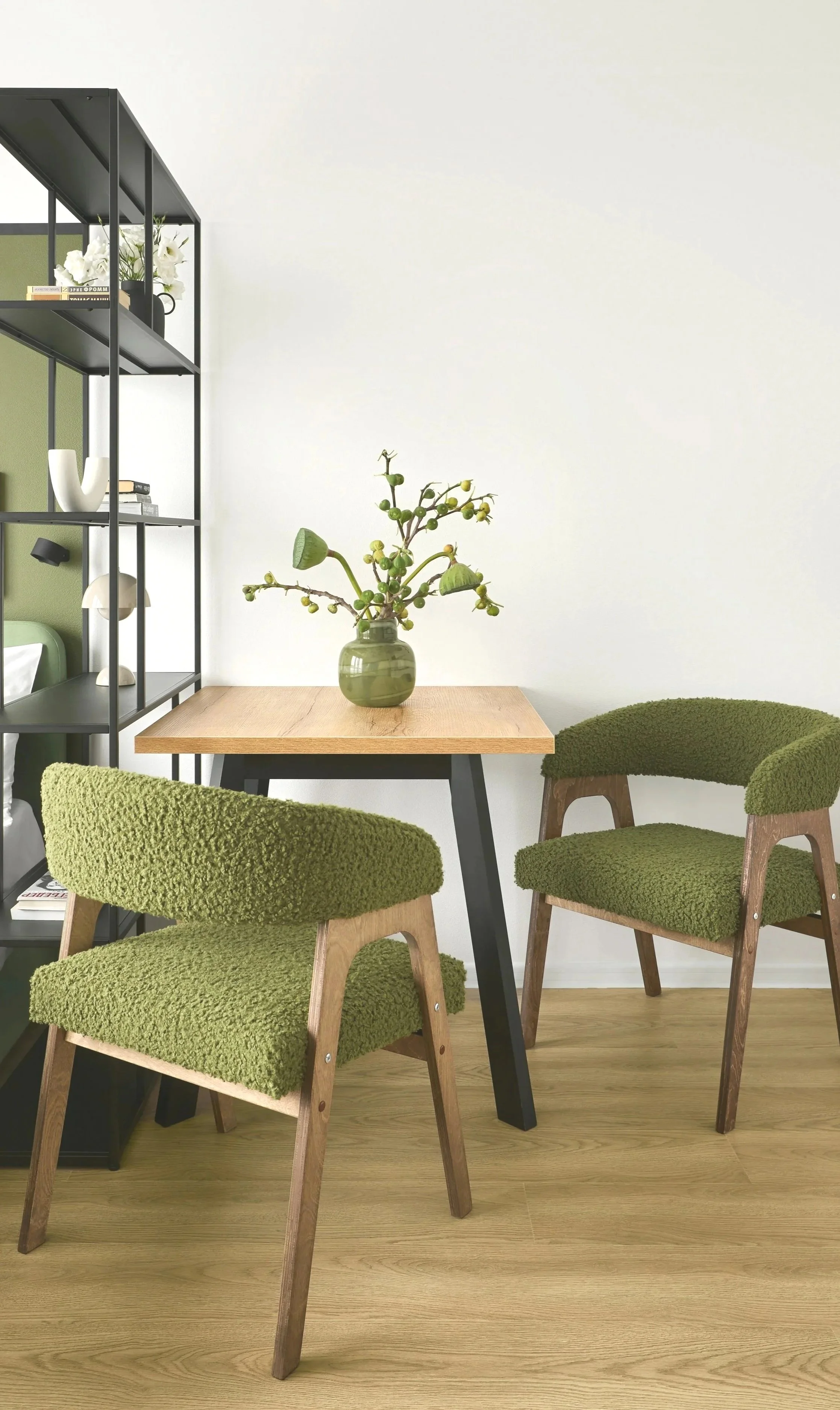 A small wooden table with a vase of plants, two green armchairs, and a black metal bookshelf against a white wall.