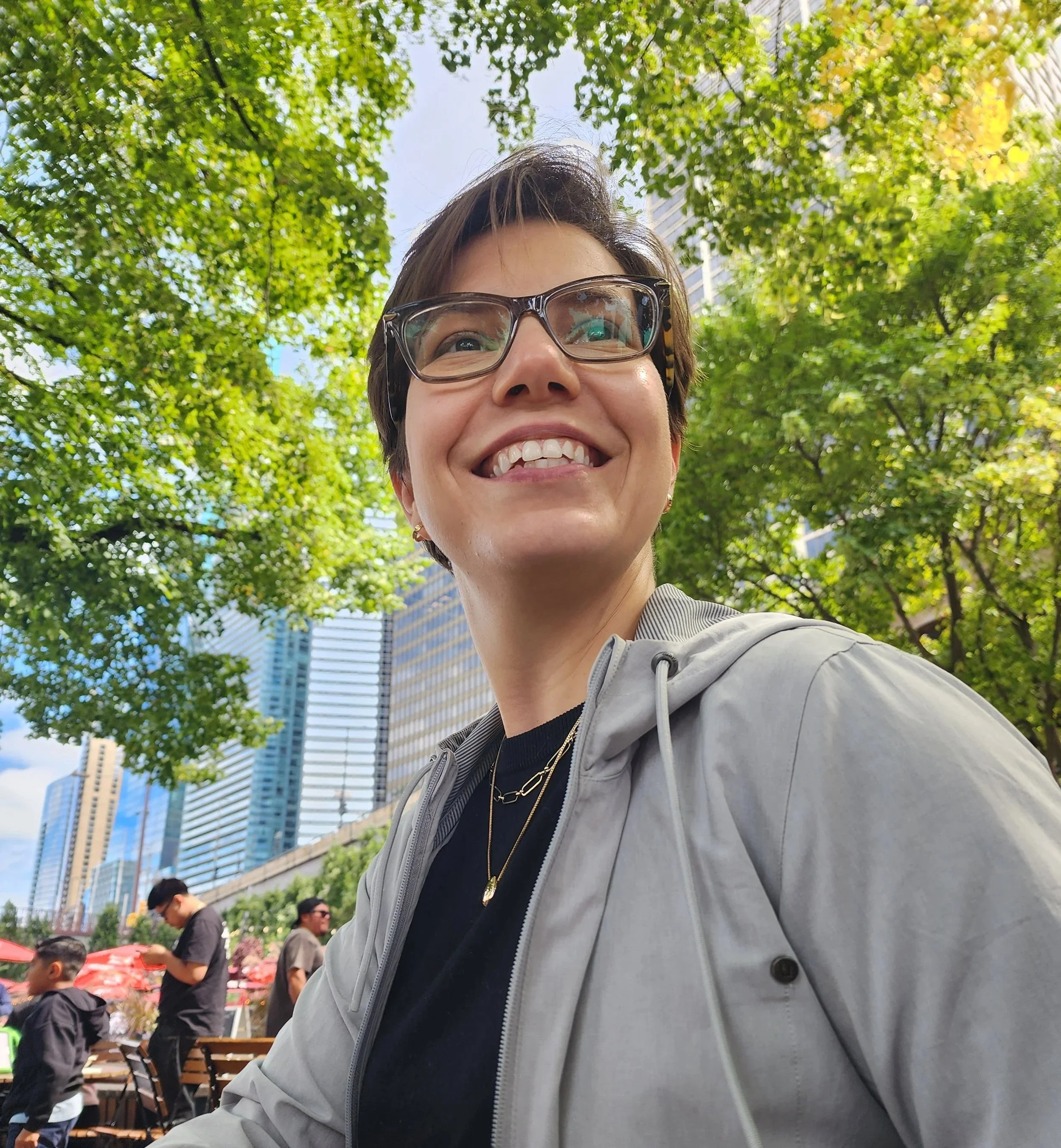 A woman smiling outdoors in a city park with tall buildings in the background. She has short brown hair, glasses, and is wearing a gray jacket, black shirt, and gold jewelry.