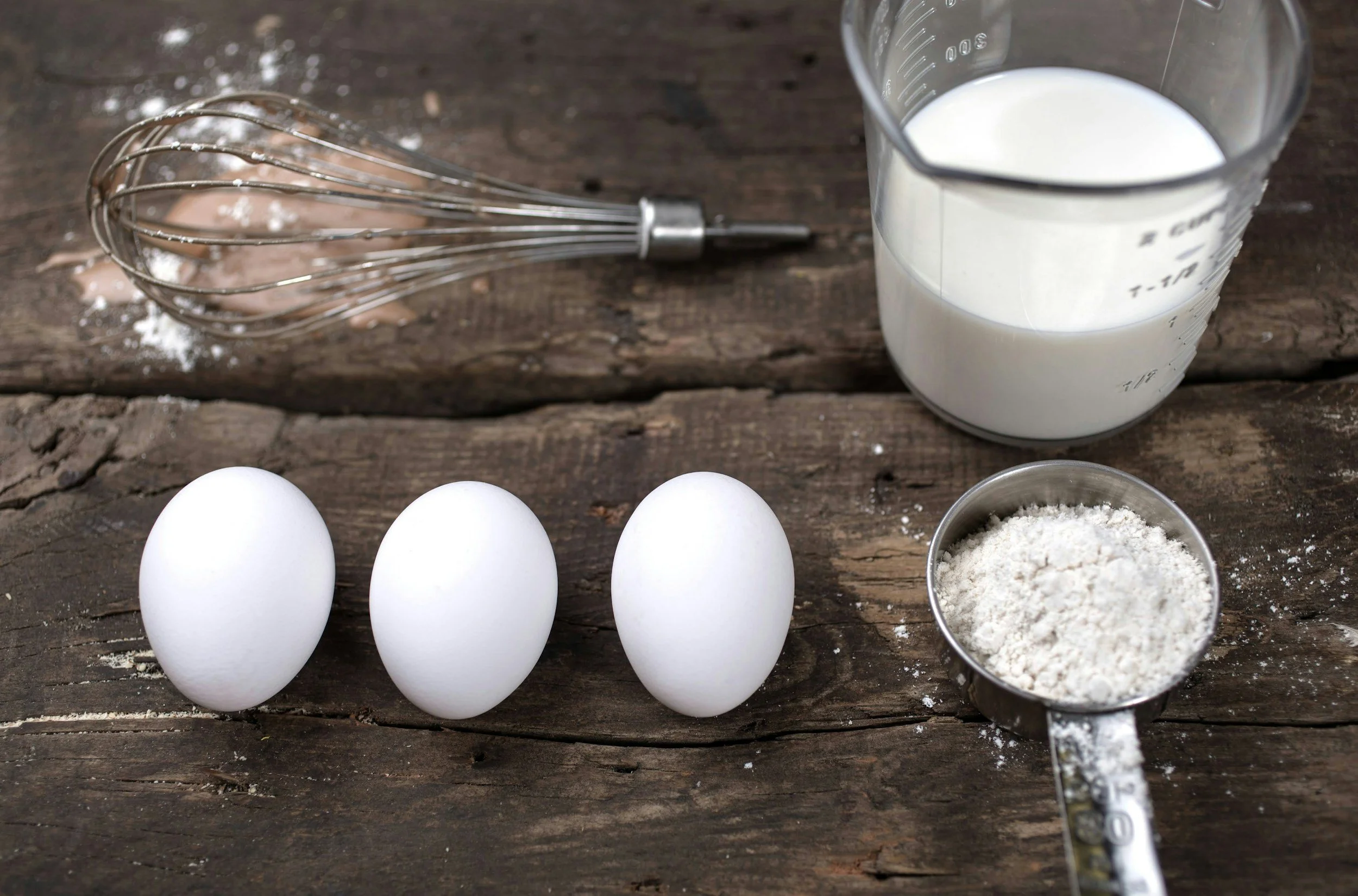 Three white eggs, a whisk with some batter or mixture, a glass measuring cup with milk, a small metal measuring cup with flour, all arranged on a rustic wooden surface.
