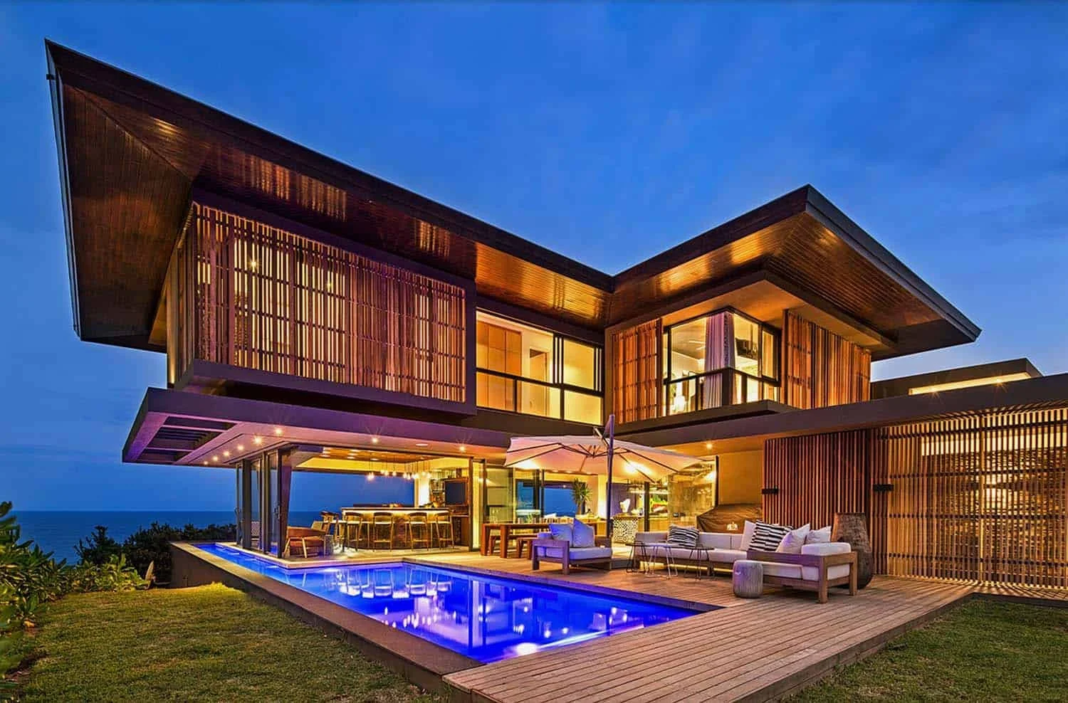 Modern two-story house with wooden exterior and large glass windows, illuminated at night, featuring an outdoor pool with blue lights, patio furniture, an umbrella, and ocean view.