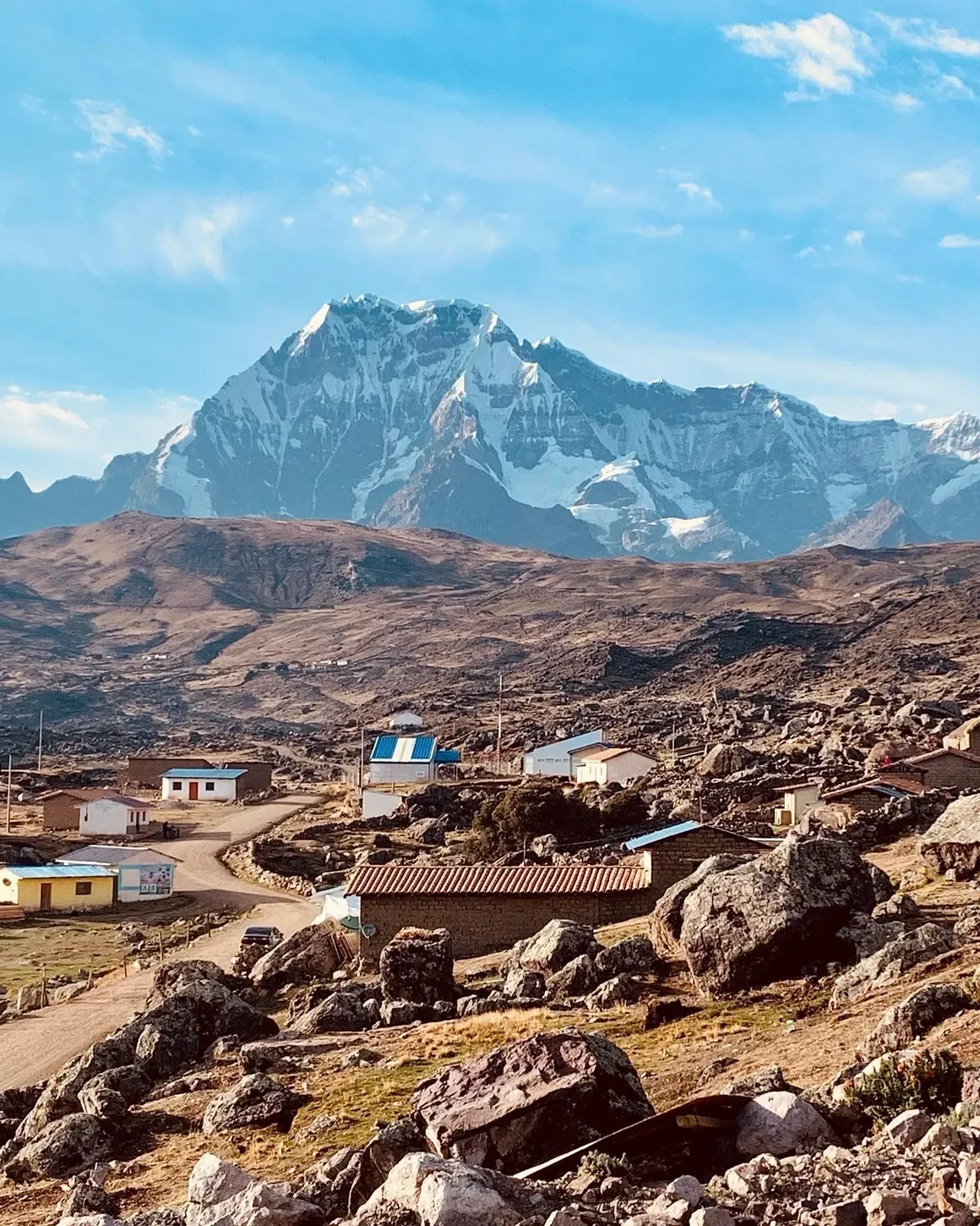 A mountain landscape with snow-capped peaks in the background, rolling brown hills, and a small village with colorful houses and dirt roads in the foreground.