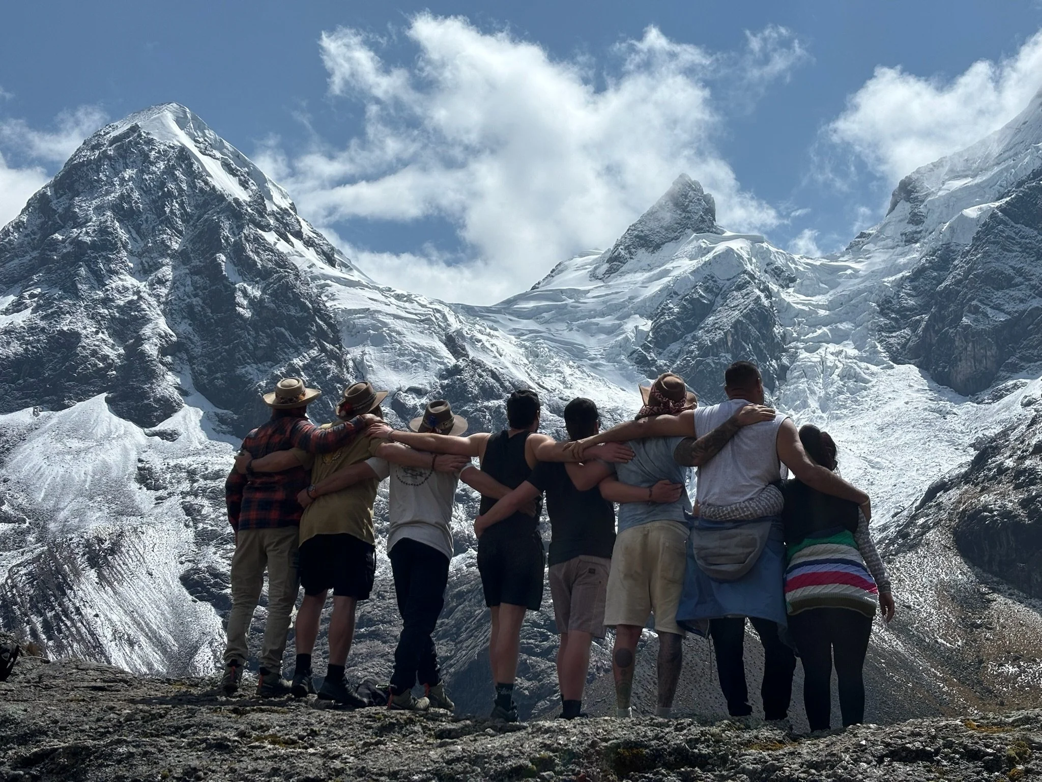 A group of people standing together with arms around each other, facing snow-covered mountain peaks on a cloudy day.