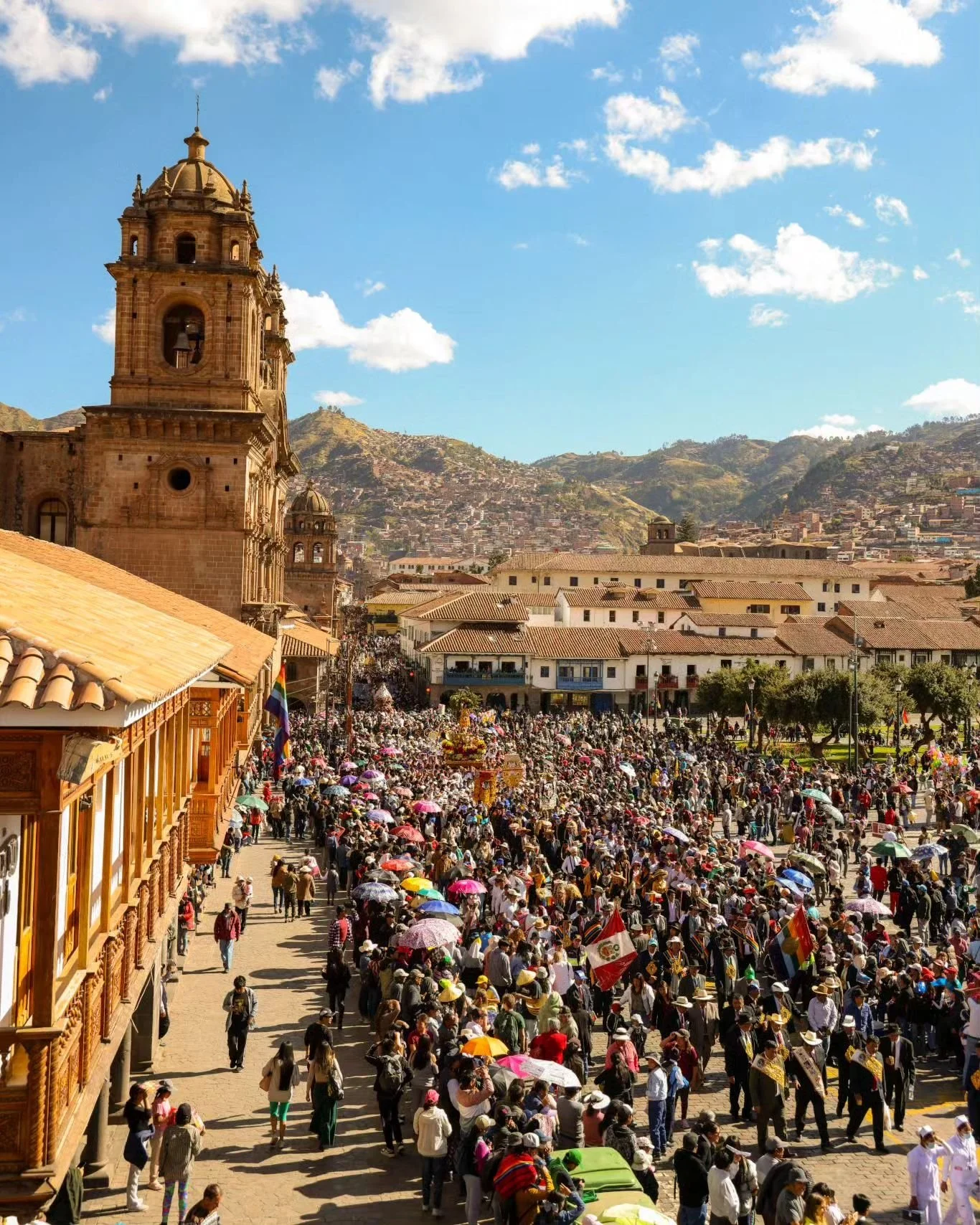 Crowd of people with umbrellas in a town square, with colonial-style architecture and a church in the background, mountains under a blue sky